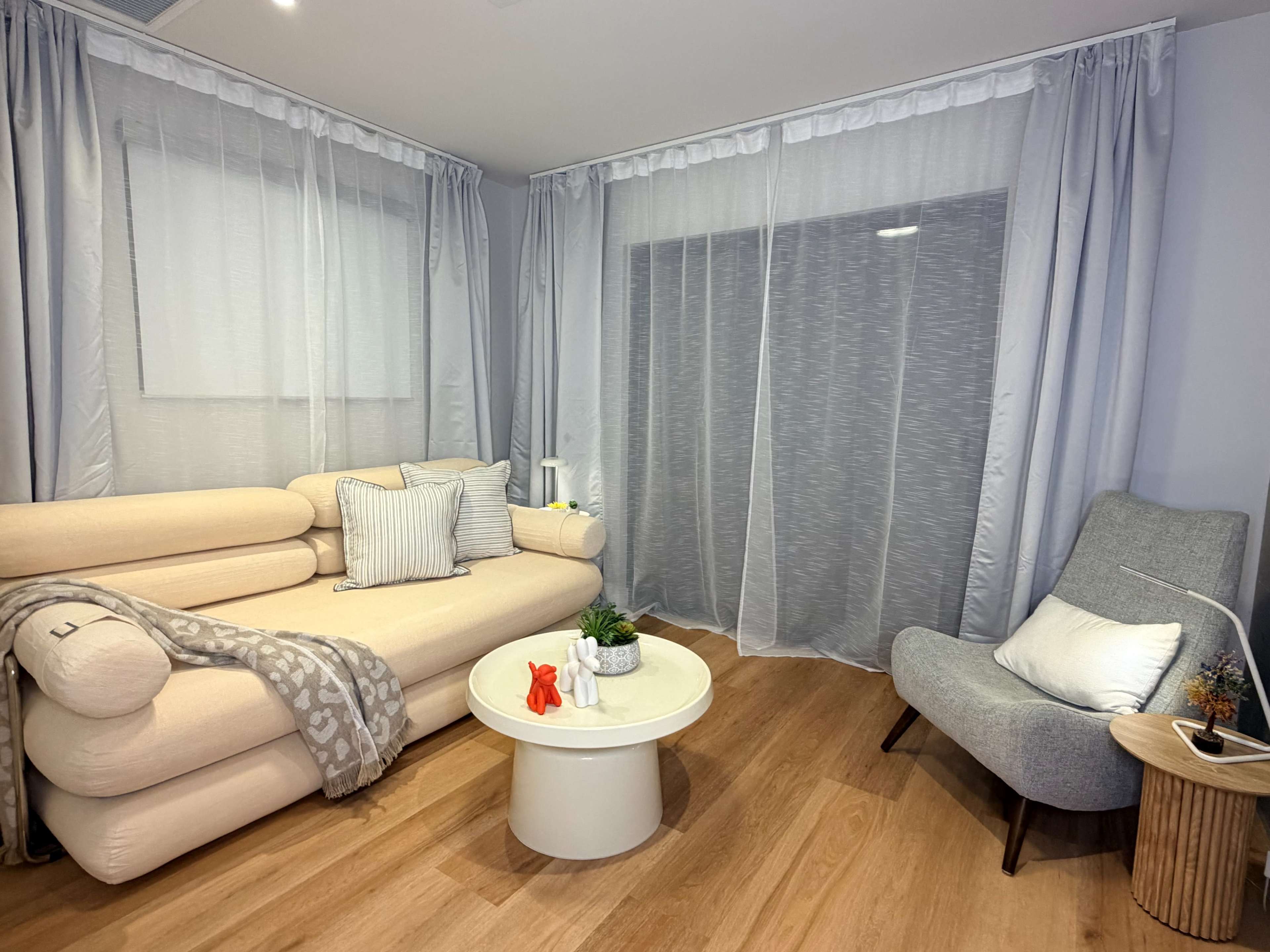 The image shows a modern living room with a light-colored sofa, a round coffee table, and sheer curtains covering the windows.