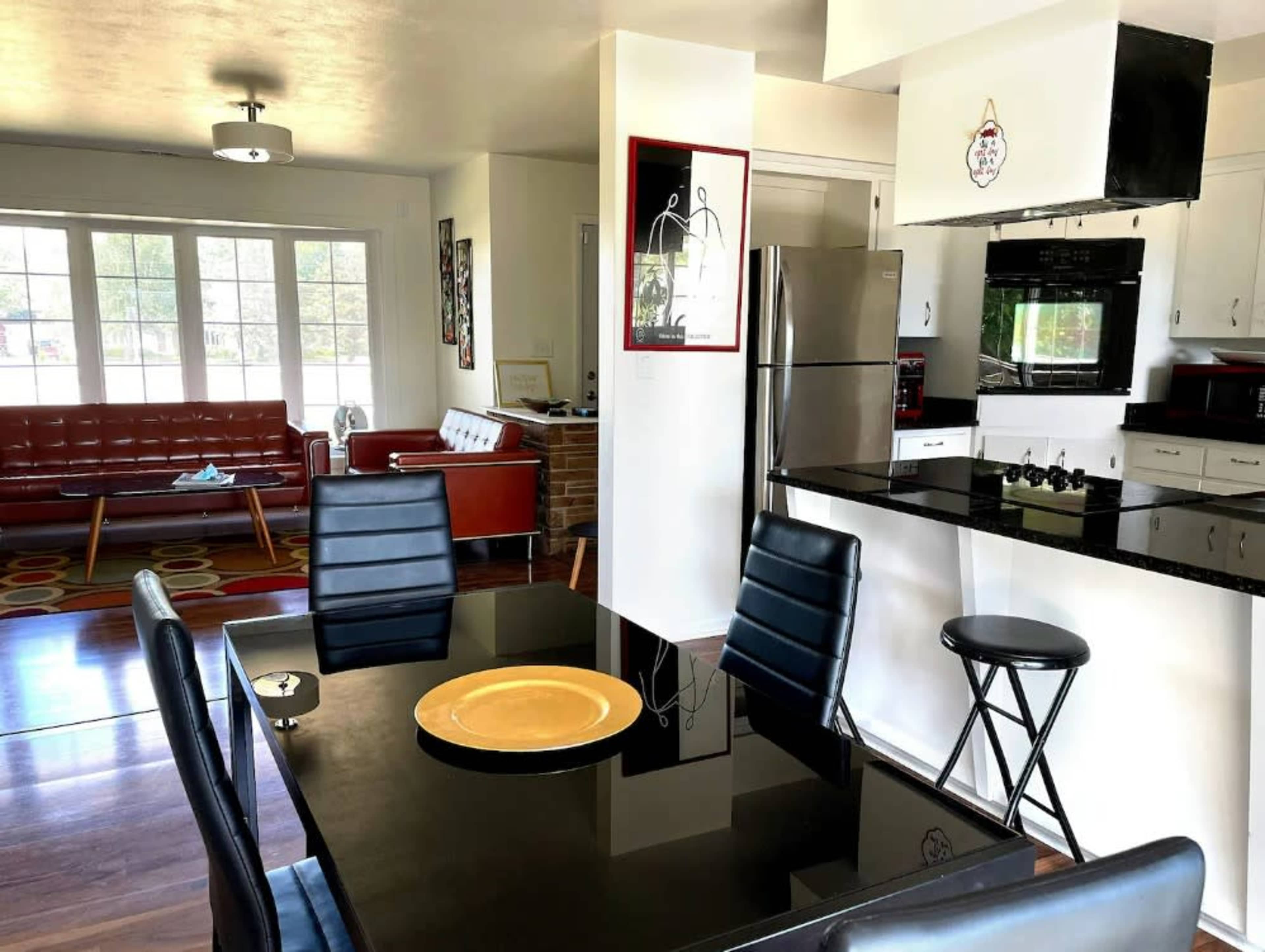 A modern open-concept living and dining area with a glass dining table and black chairs, adjacent to a kitchen featuring stainless steel appliances and a red sofa in the background.