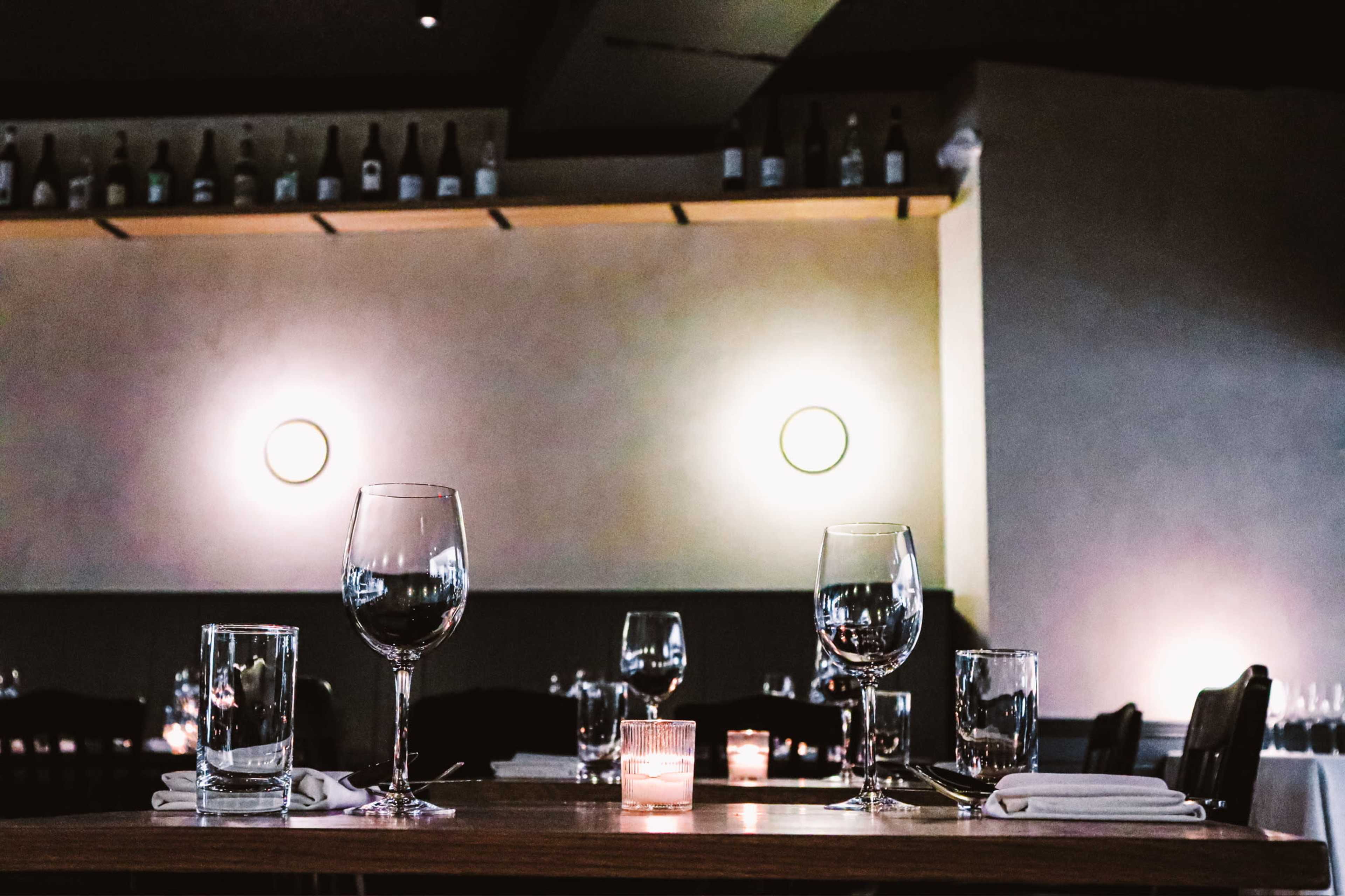 A dining table is set with wine glasses, water glasses, and a lit candle in a restaurant with dim lighting and bottles on a shelf in the background.