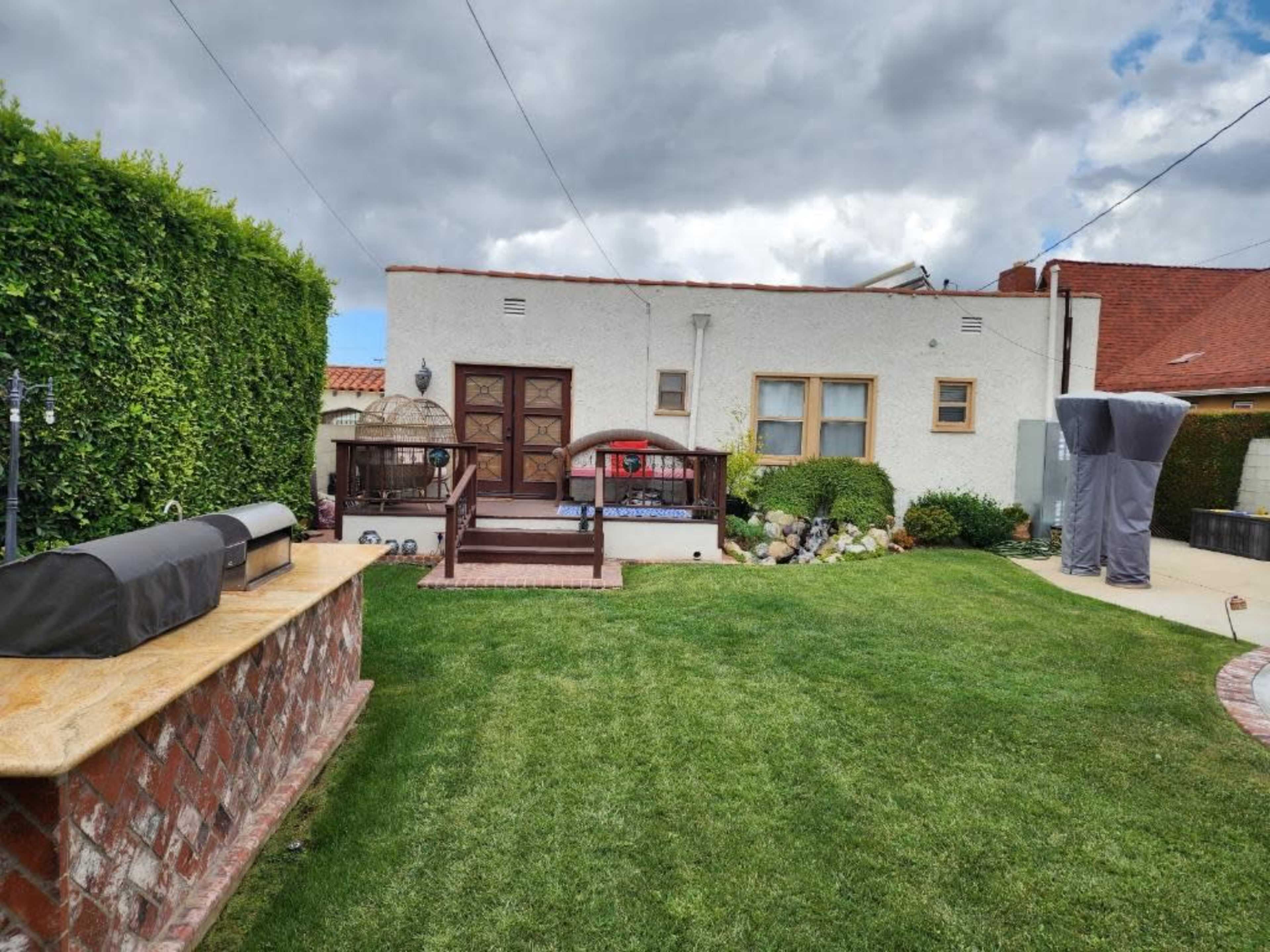 The image shows a backyard with a green lawn, a wooden deck leading to a light-colored house, and a brick outdoor fireplace beside a grill setup.