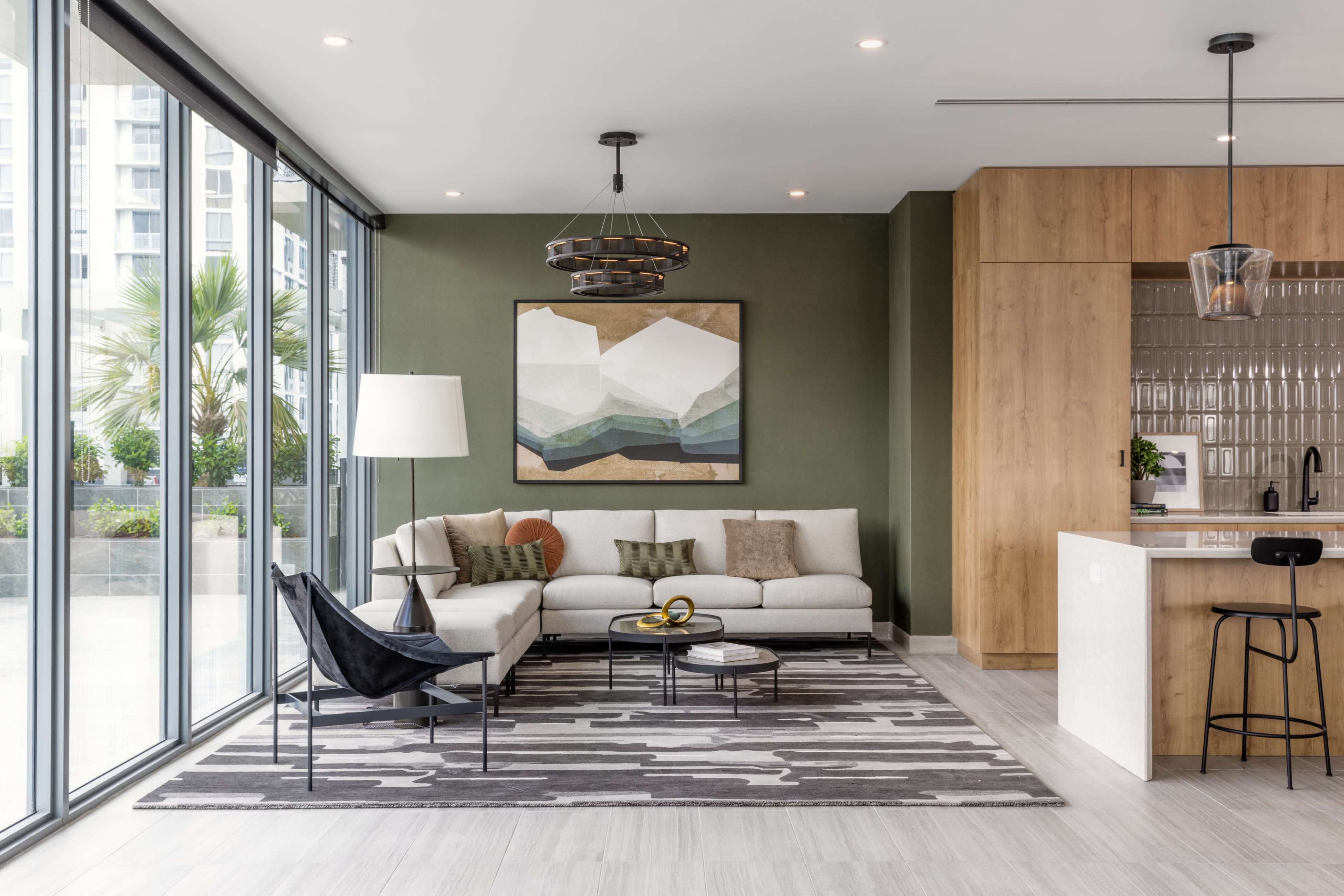 A modern living room with large windows, featuring a light-colored sofa, a geometric wall art piece, a round coffee table, and a kitchen area in the background.