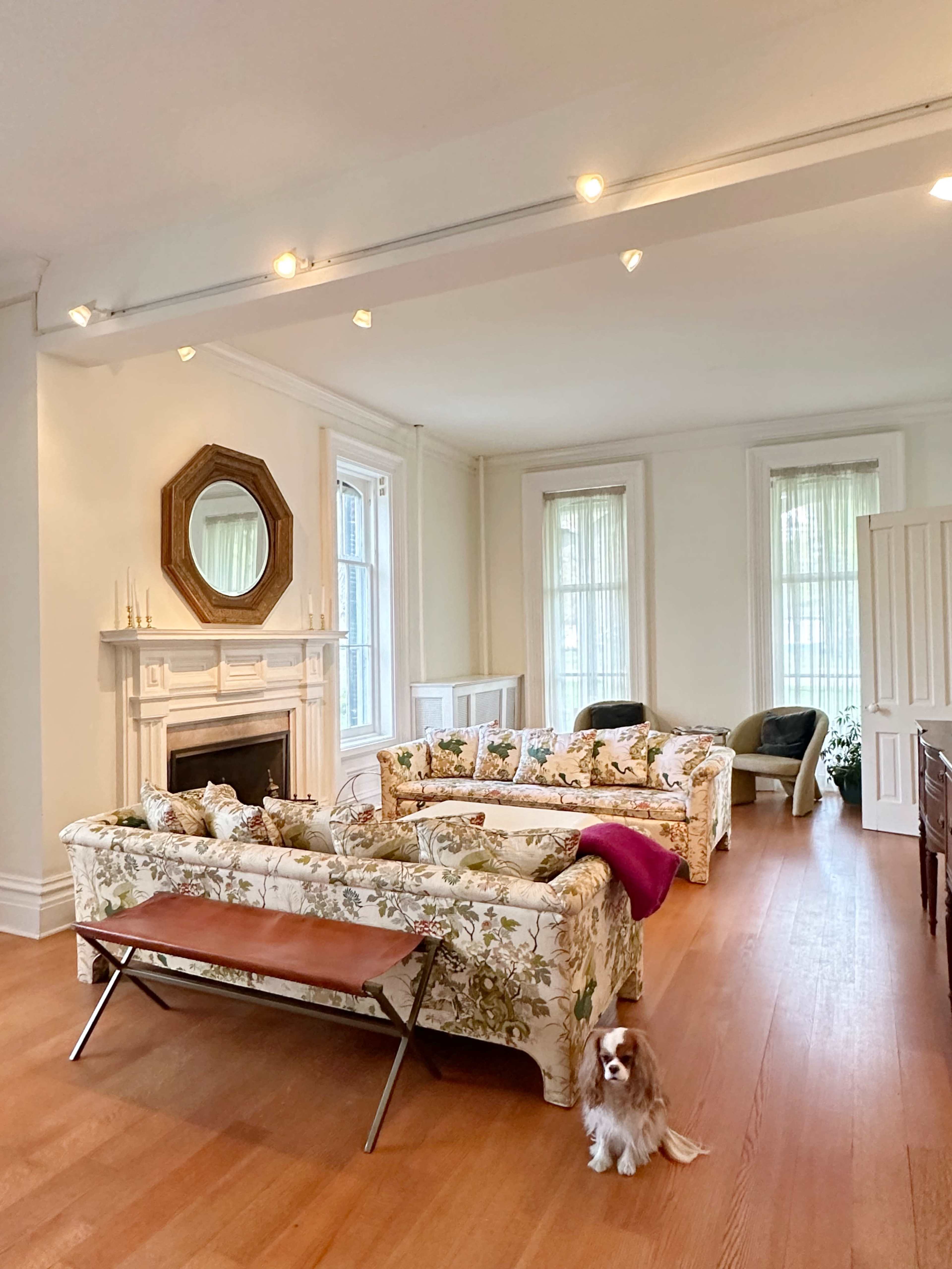 A spacious living room with floral-patterned couches, a coffee table, a fireplace, and a small dog on the wooden floor.