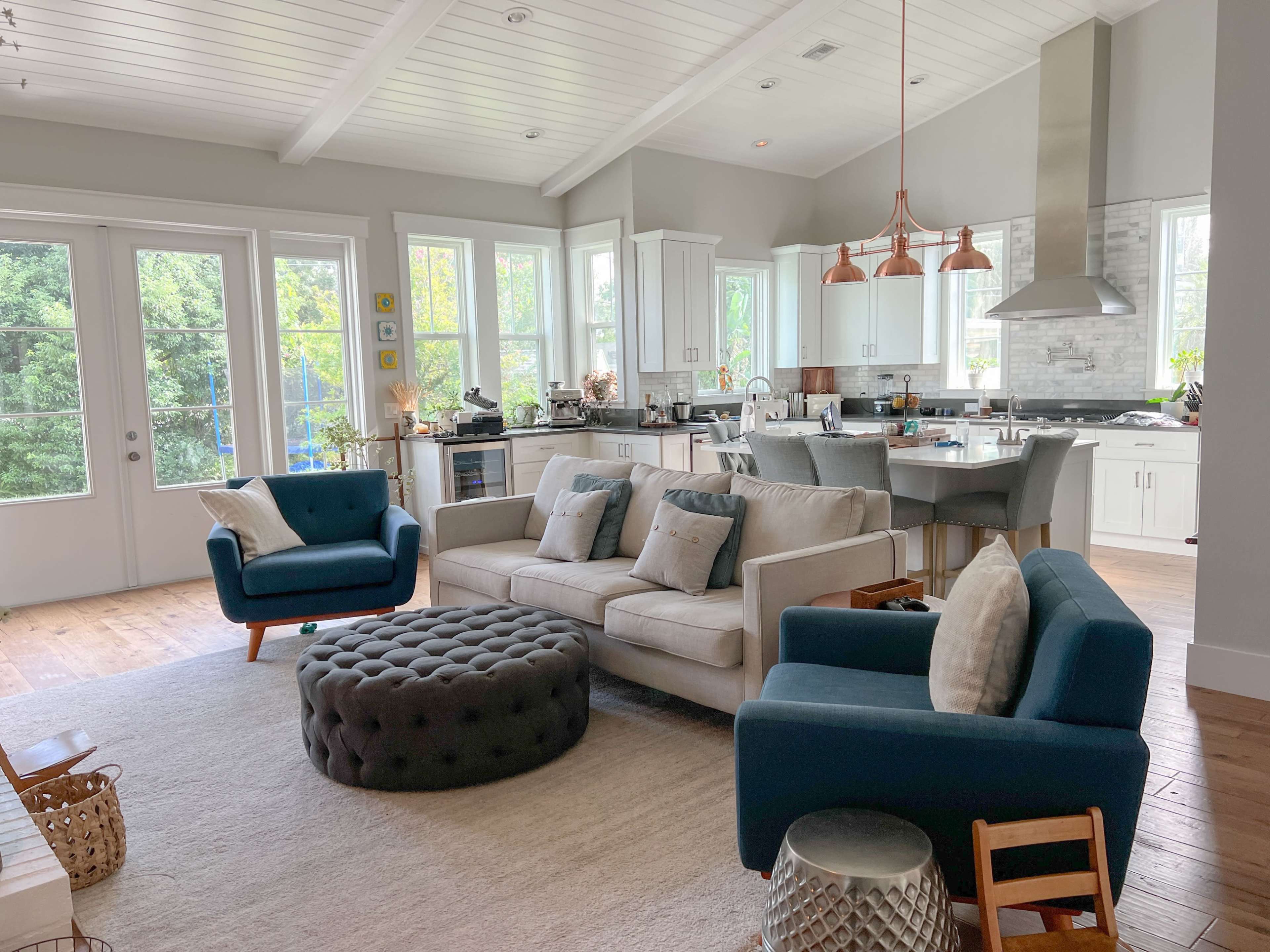 A bright, modern living room features a light-colored sofa, two dark blue chairs, a tufted ottoman, and a kitchen area with white cabinets and metallic accents.