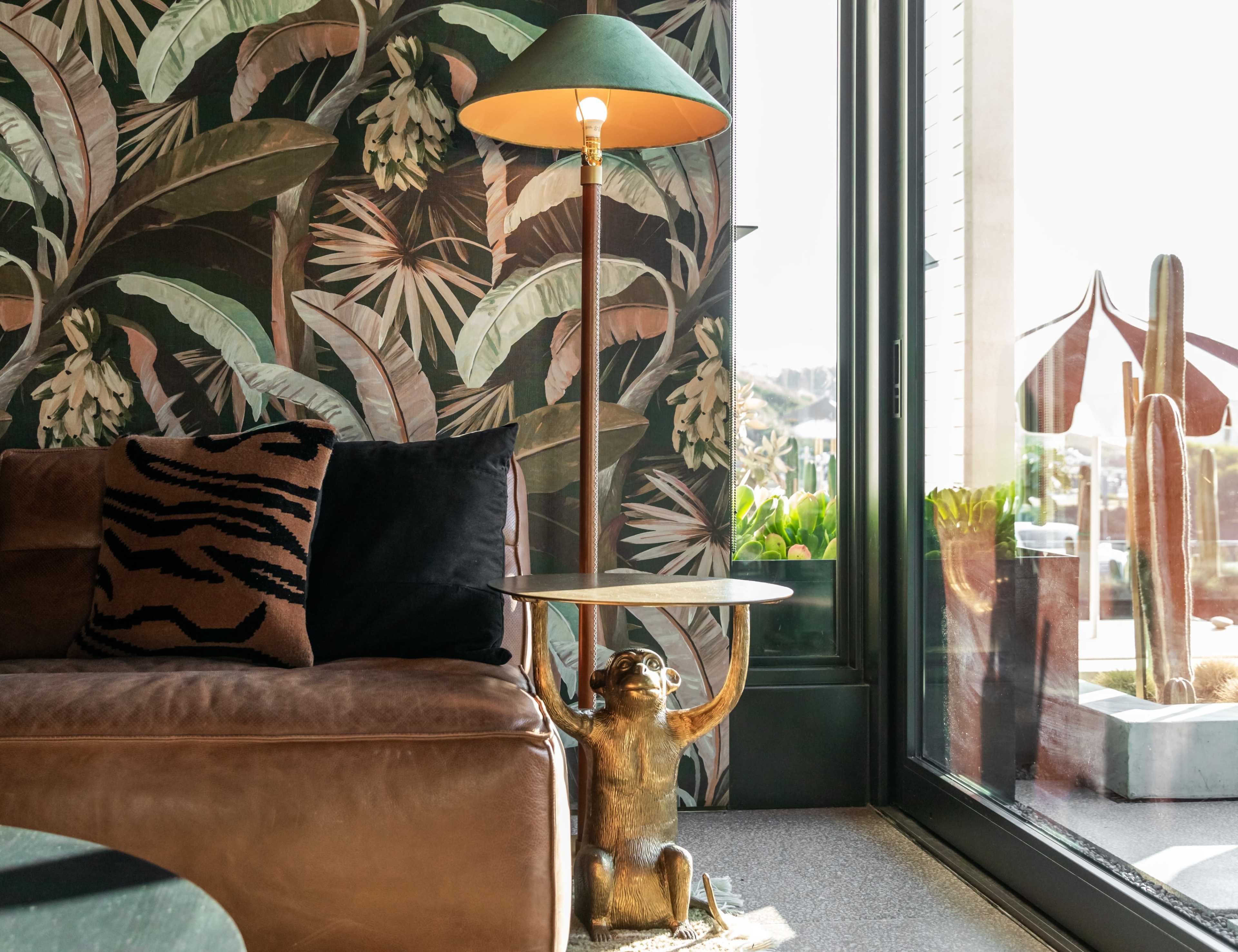 A living room features a brown leather sofa, a decorative monkey lamp with a green shade, and a large window overlooking a garden with cacti.