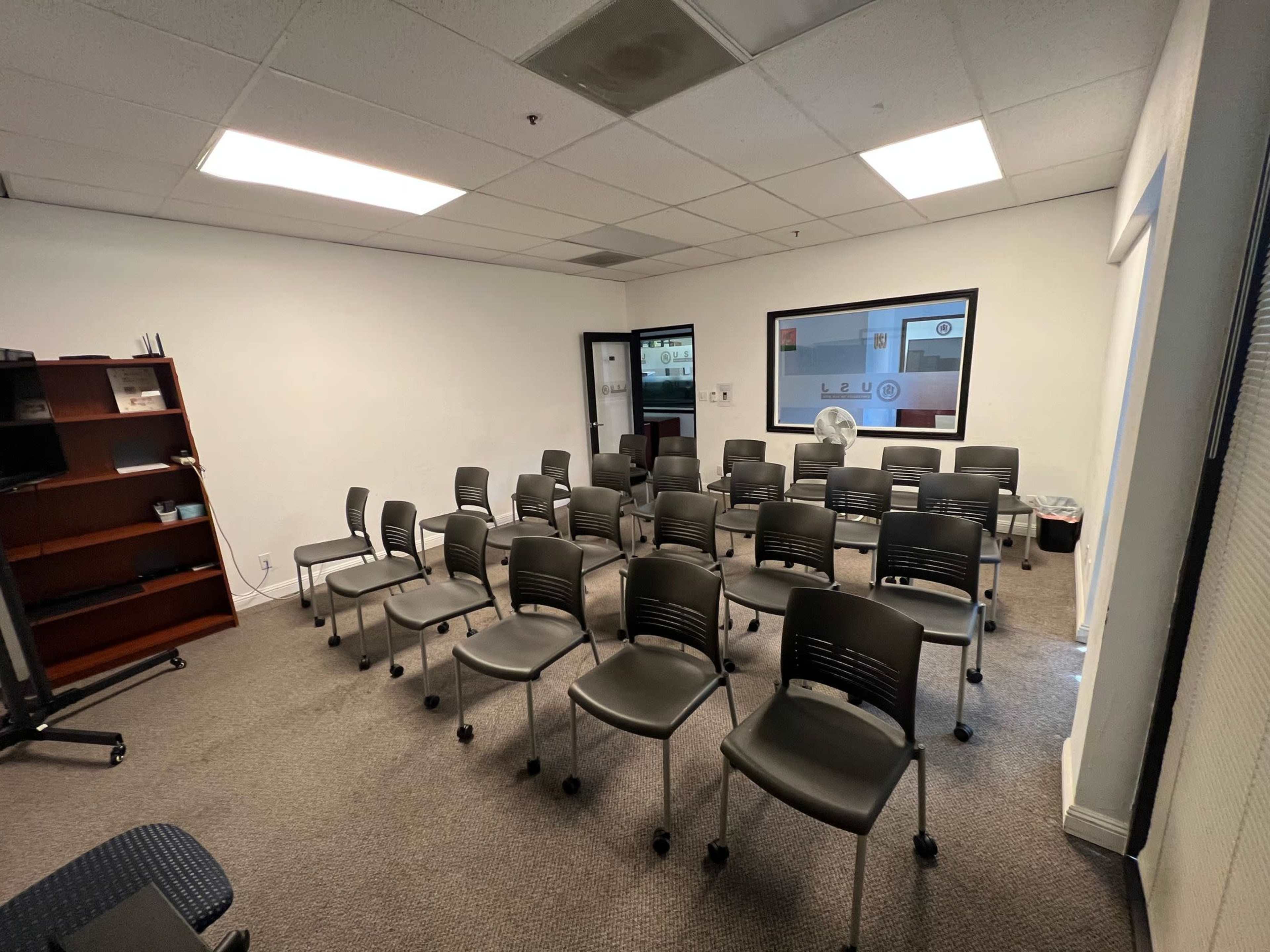 A sparsely furnished meeting room with rows of gray chairs arranged in a semi-circle facing a whiteboard or presentation screen.