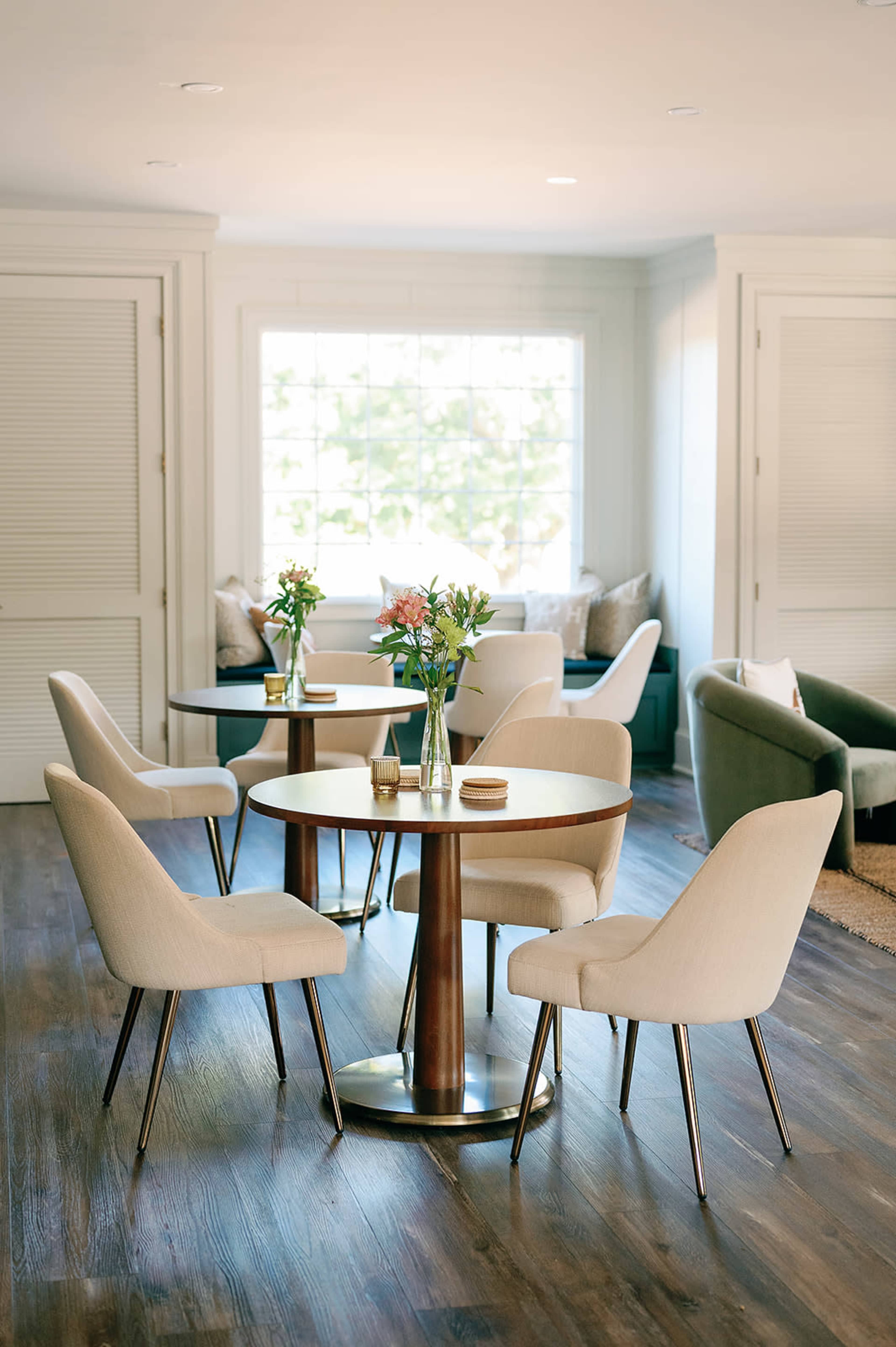 A round table surrounded by five light-colored chairs is situated in a well-lit room with large windows and a cozy seating area.