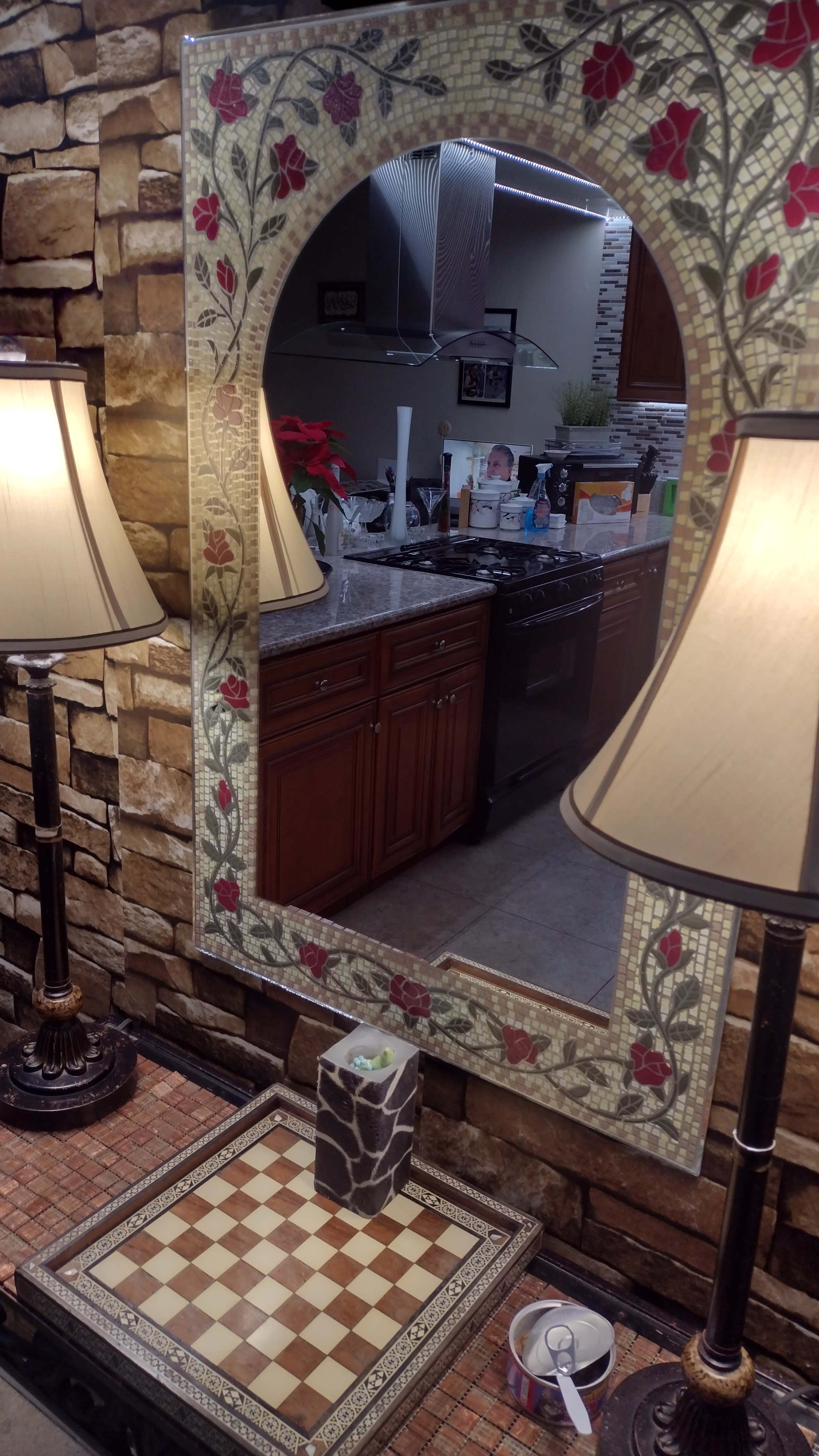 A large, decorative mirror with floral motifs reflecting a kitchen featuring wooden cabinets and a stove, alongside two lamps on a checkered base.