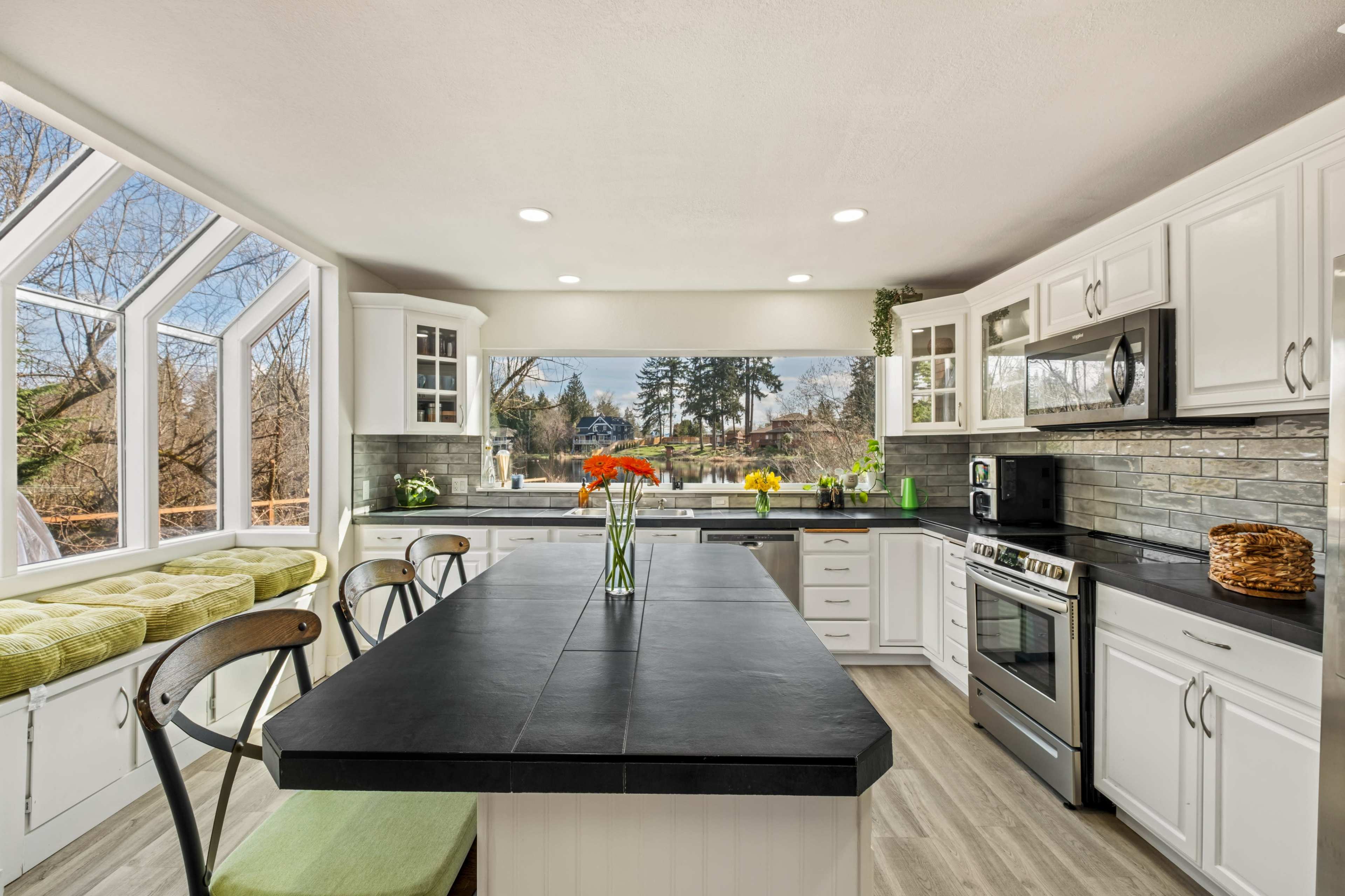 A spacious kitchen features a large black island countertop, white cabinetry, and a windowed area with a cushioned seating space overlooking the outdoors.