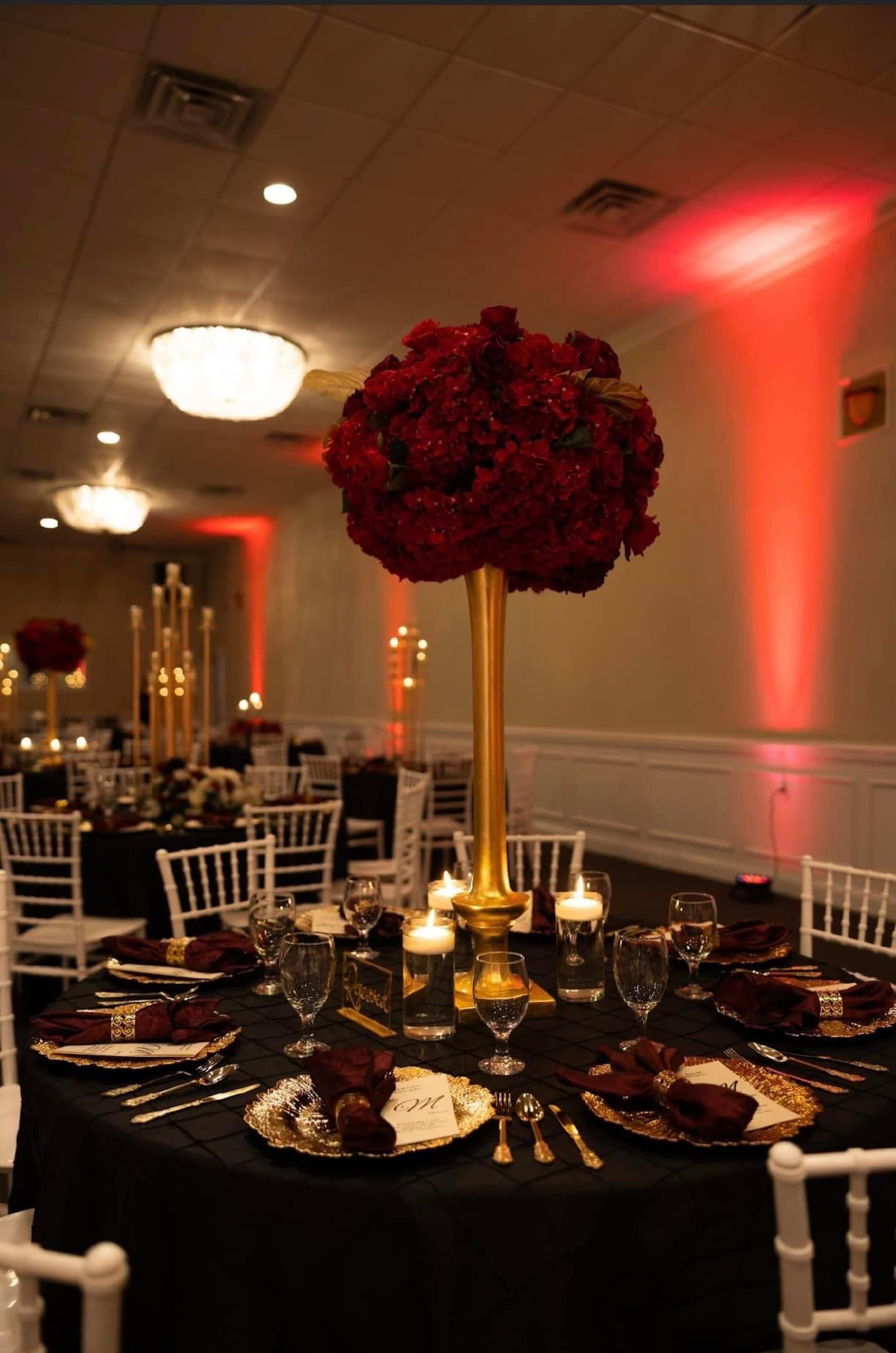 A banquet hall is elegantly decorated with tall floral centerpieces, black and gold table settings, and ambient lighting.