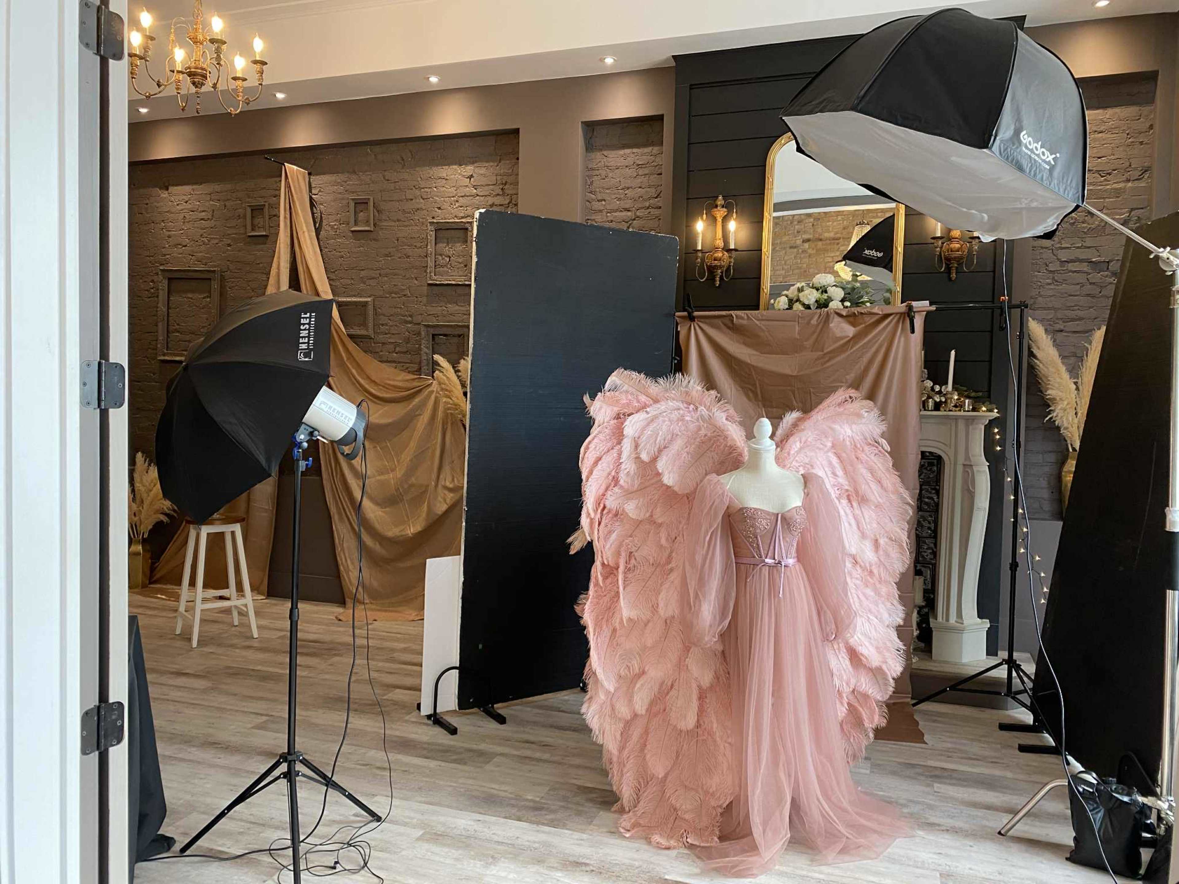 A mannequin dressed in a pink, feathered gown stands in a well-decorated photography studio, surrounded by backdrops and lighting equipment.