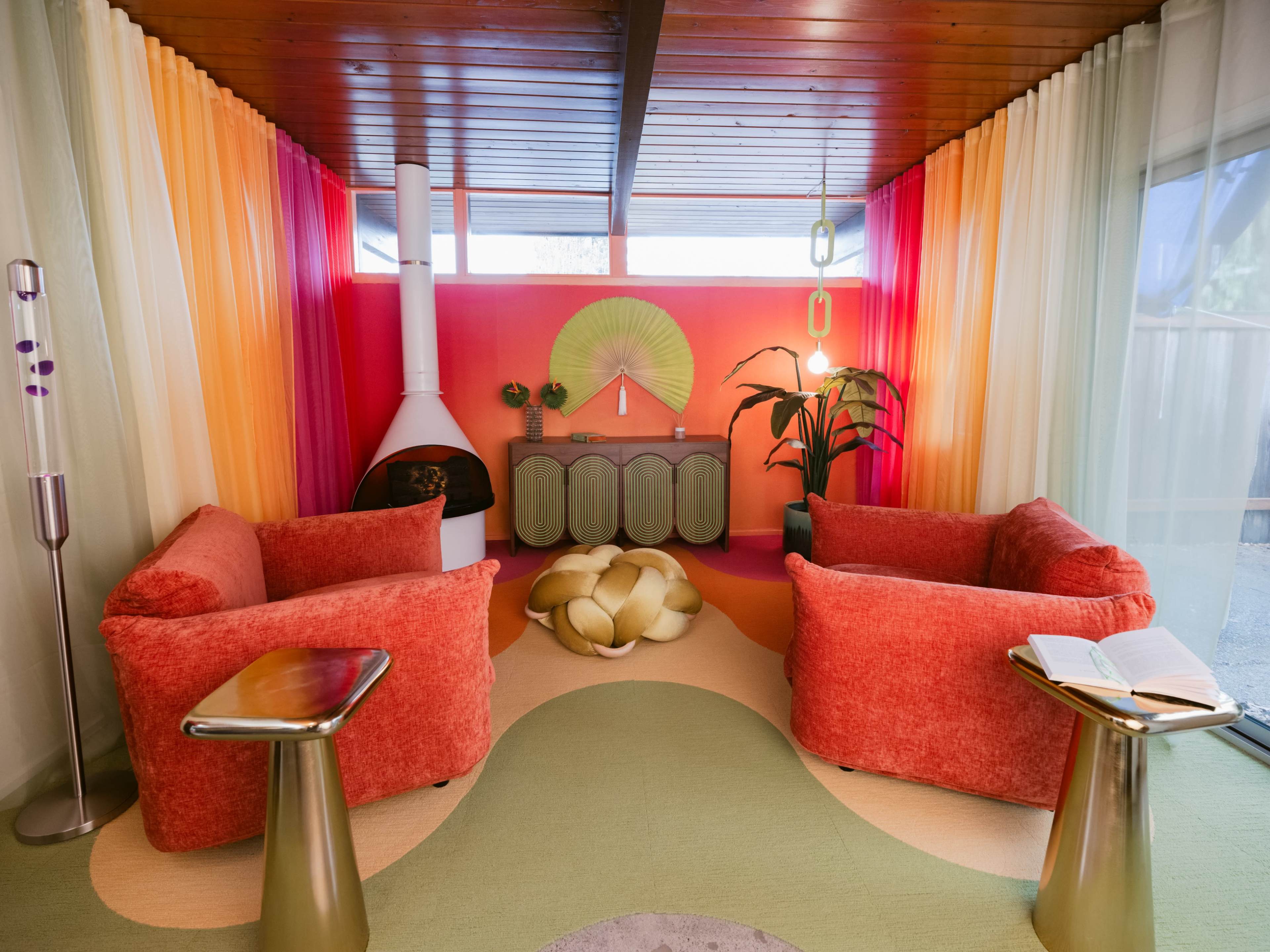The image shows a colorful living room with two red sofas facing each other, a unique circular coffee table, a decorative wall feature, and a green plant beside a modern fireplace.