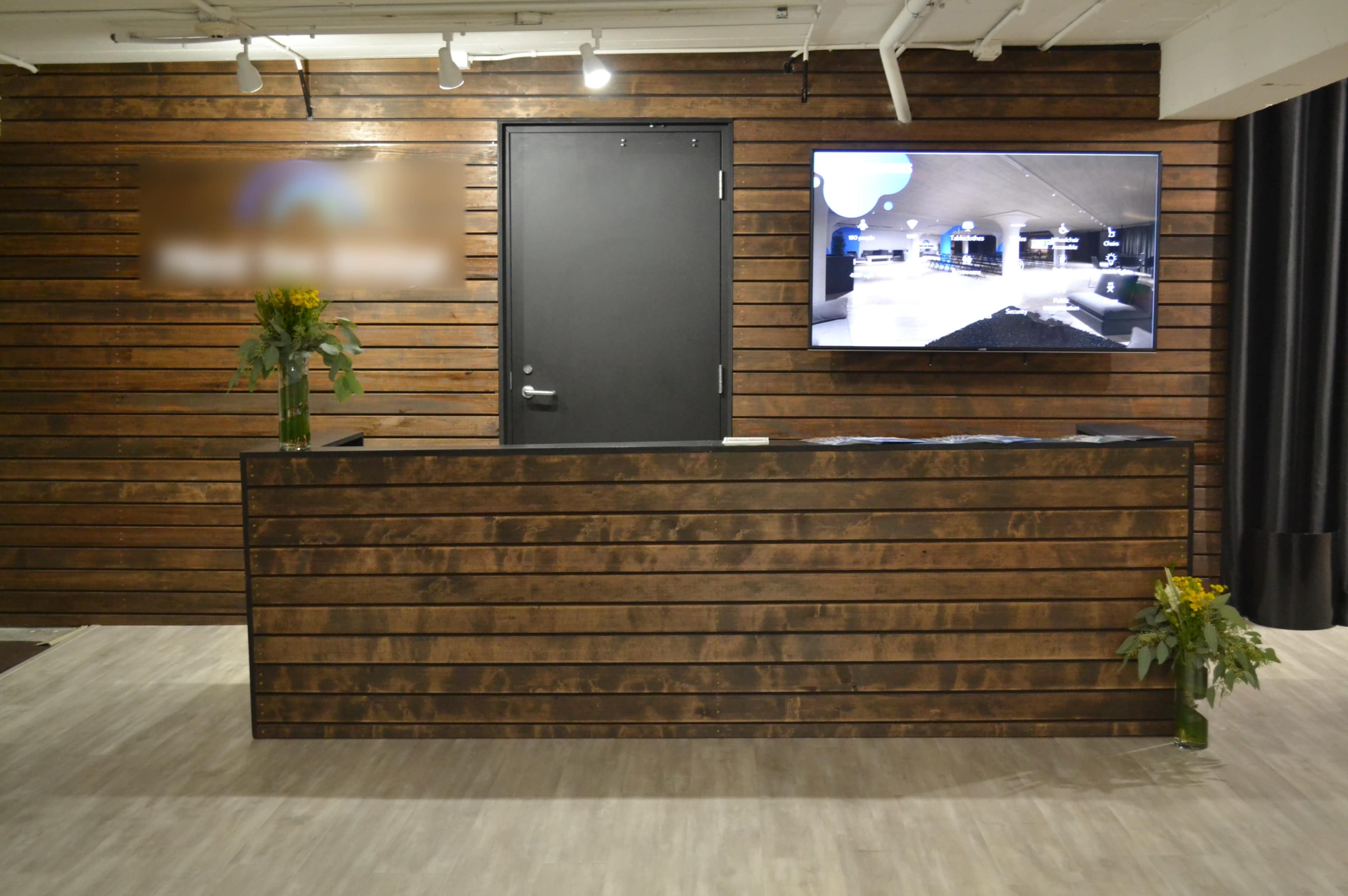 A reception area with a dark wooden counter, a large television mounted on the wall displaying a screen, and a door behind the counter.