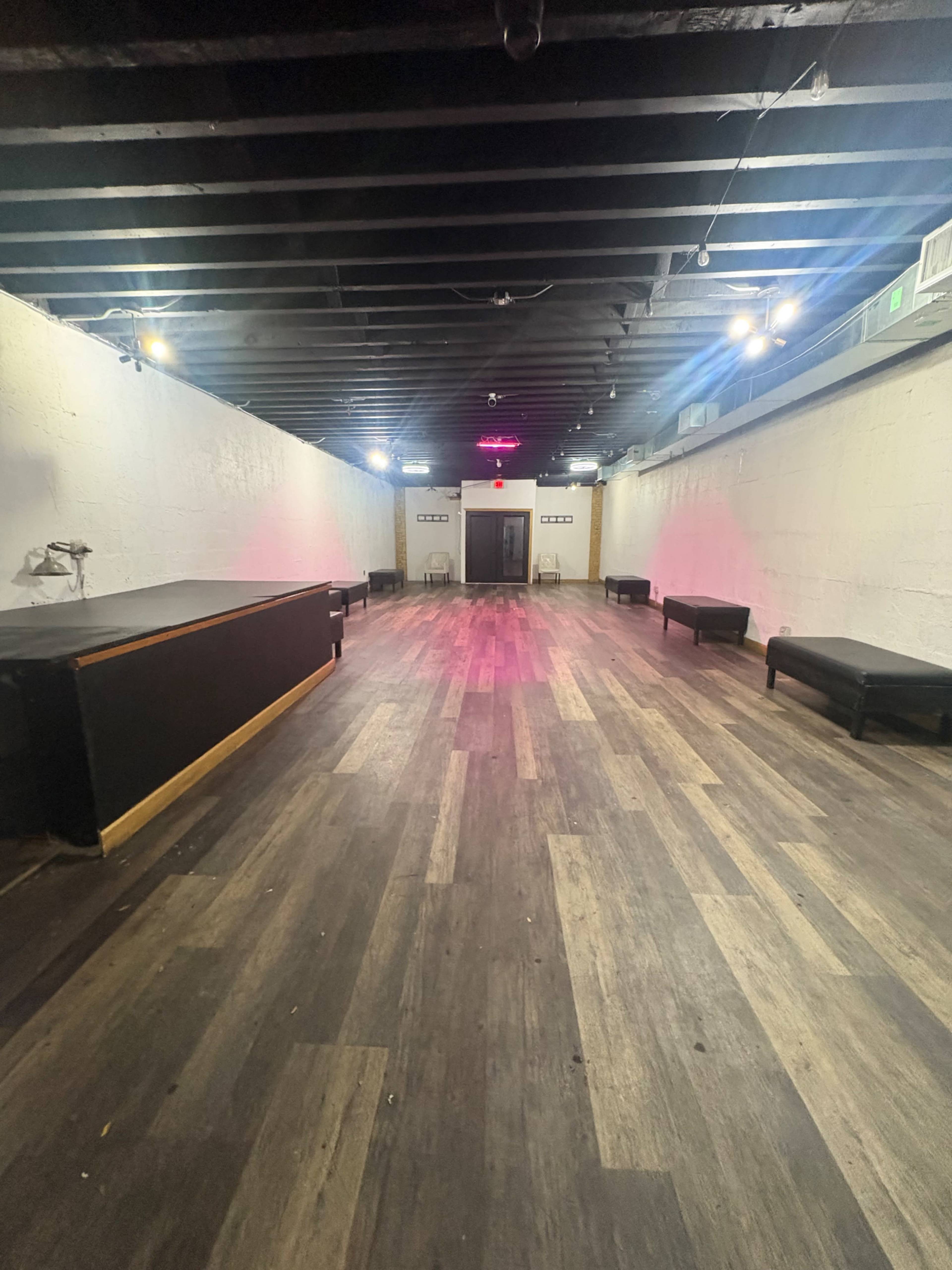 The image shows a long, empty room with wooden flooring, black benches along the sides, and a small stage or counter at one end.