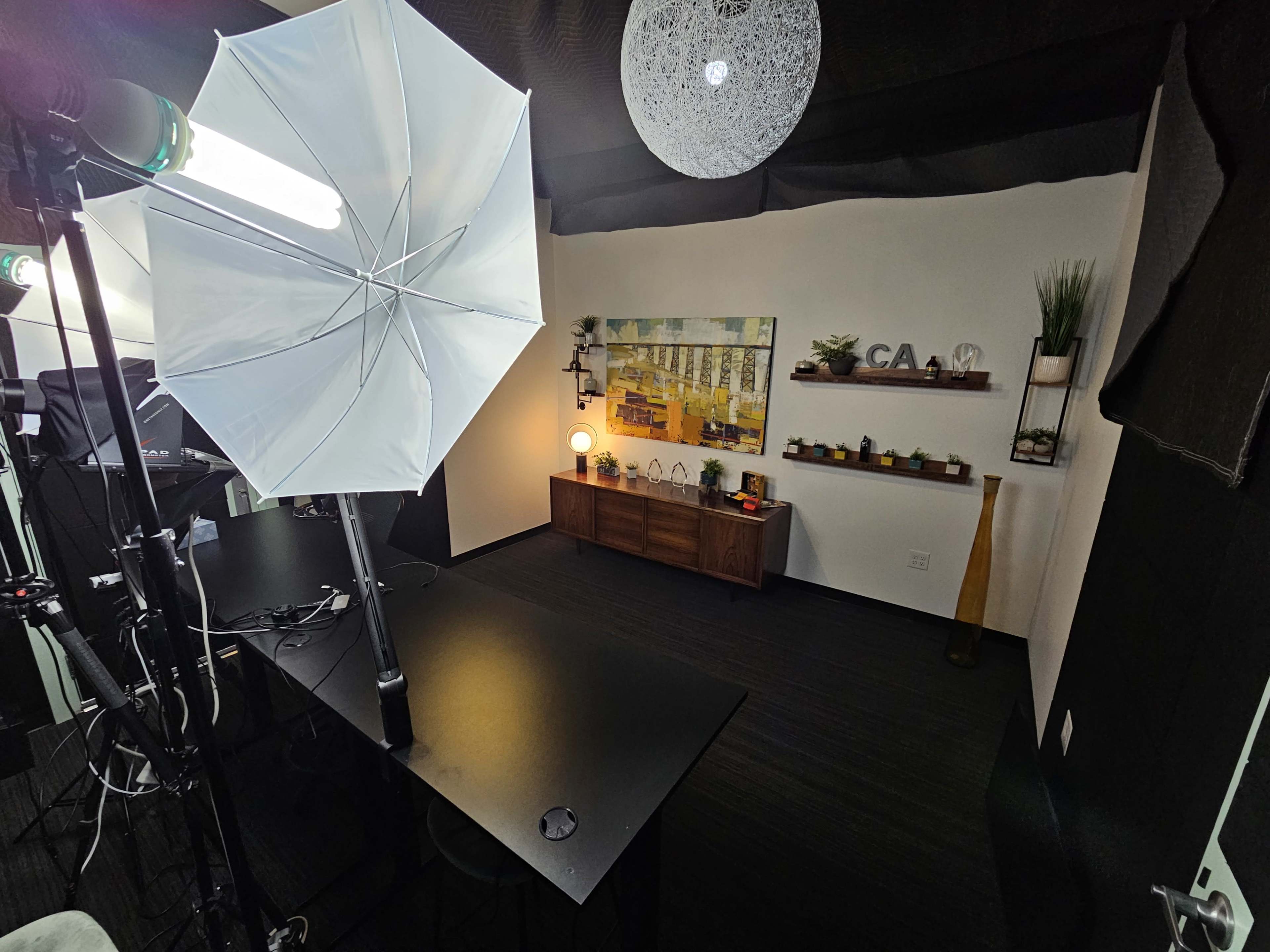 A well-lit studio space with a large umbrella light, a dark table, and decorative shelves on a white wall.