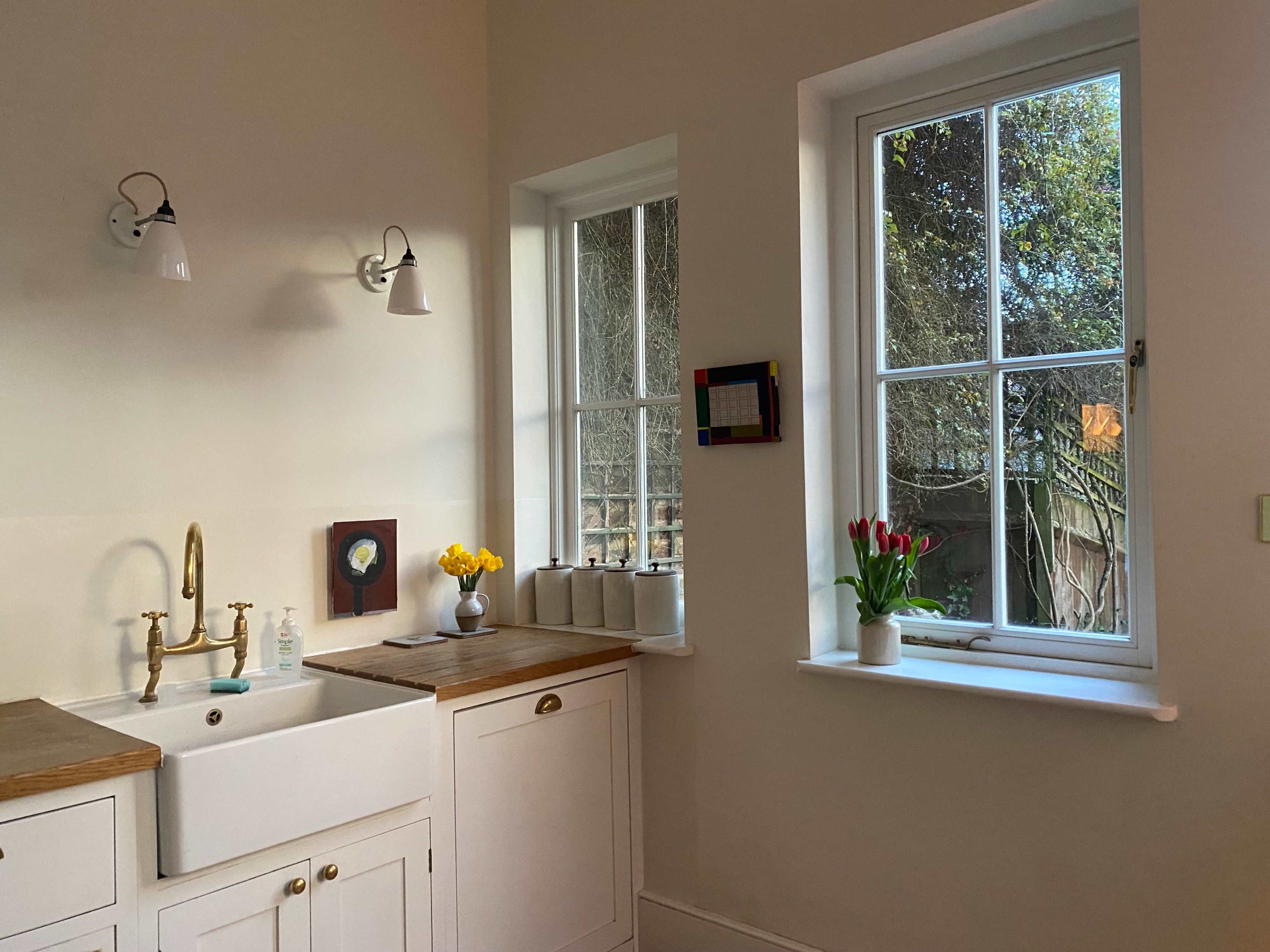 A modern kitchen with a farmhouse sink, wood countertops, and large windows letting in natural light, adorned with a vase of tulips and a colorful wall hanging.