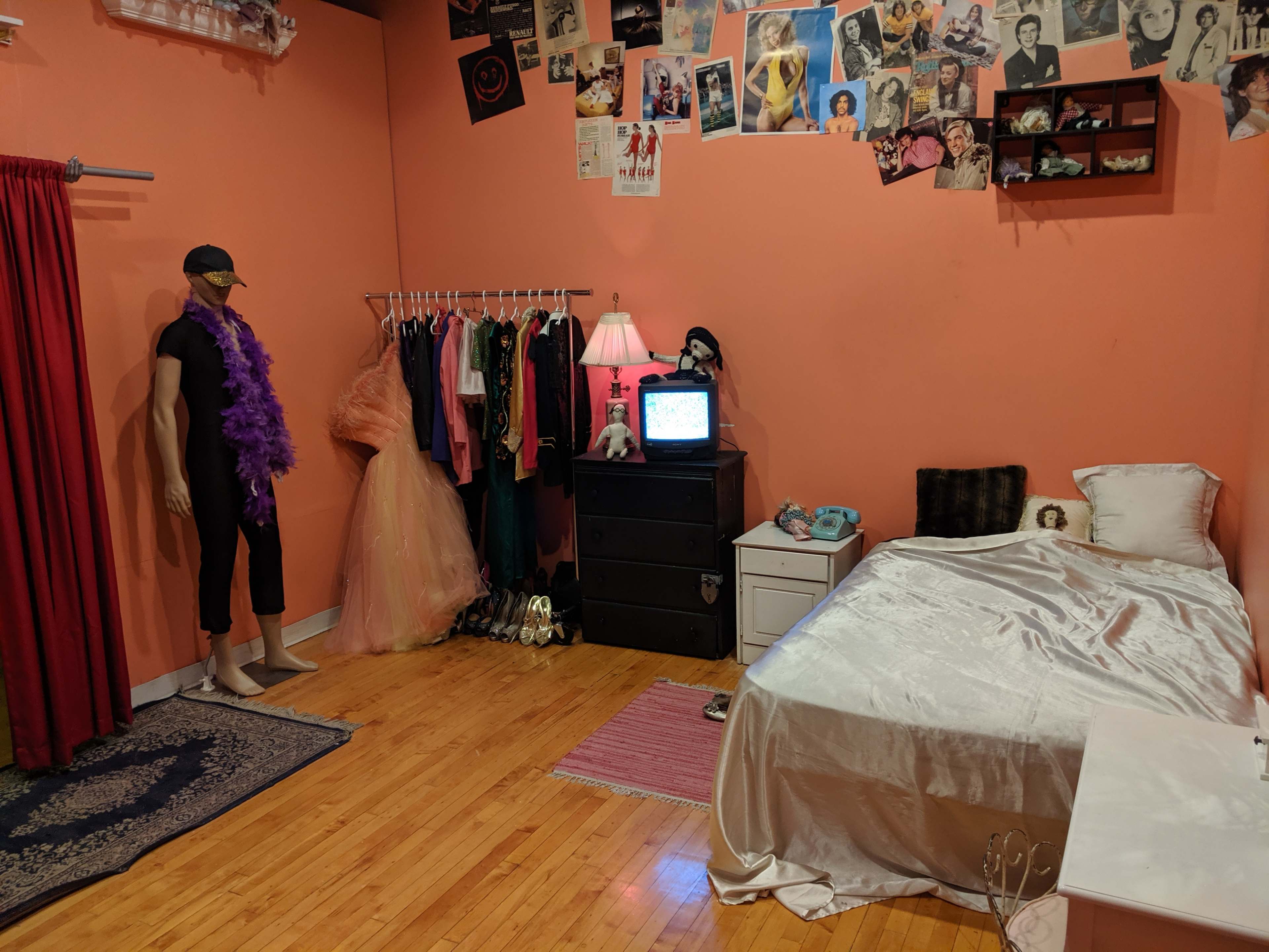 A room with an orange wall, a bed with white bedding, a mannequin dressed in black near a rack of costumes, a dresser, and a vintage television on a table.