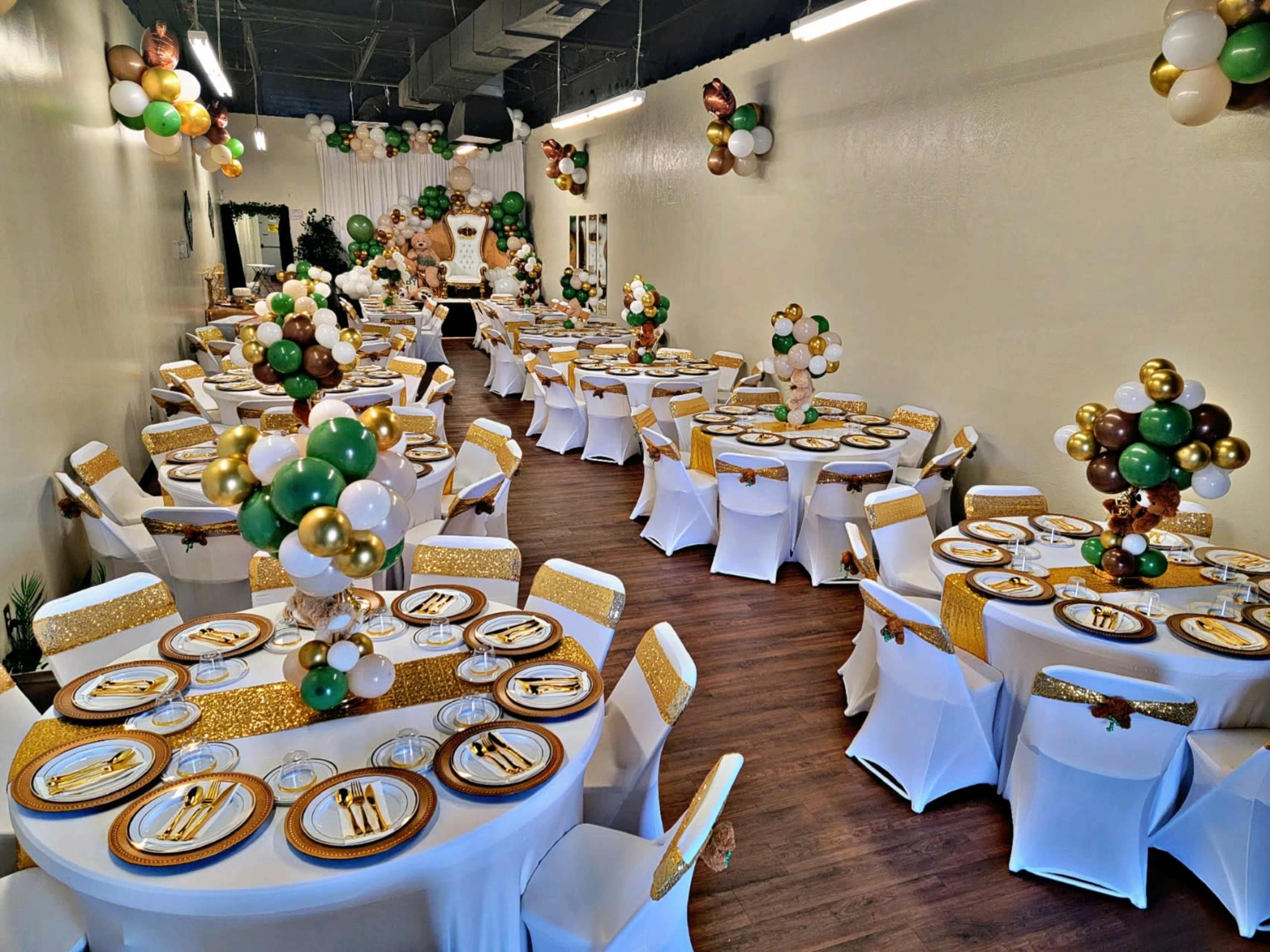 The image shows a banquet hall set up for an event, featuring multiple decorated tables with white tablecloths and various arrangements of balloons and centerpieces.