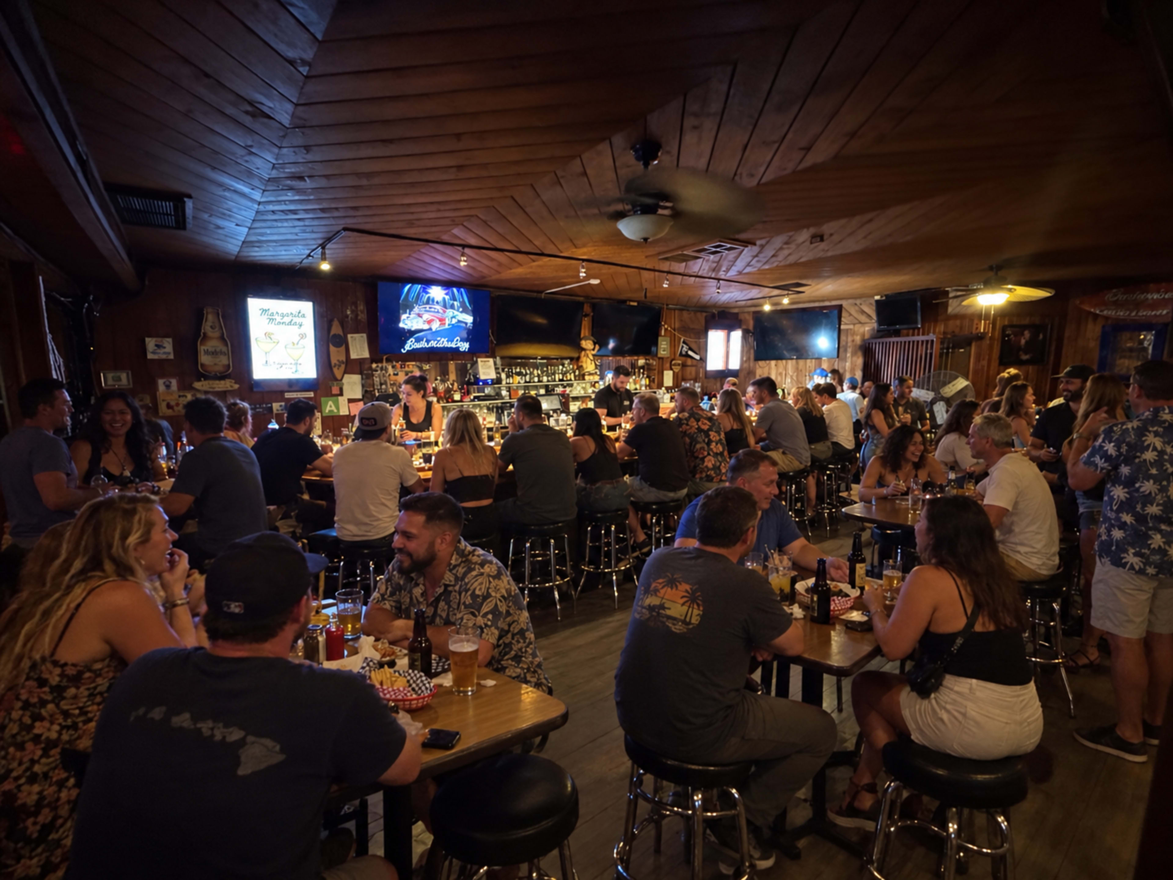 A crowded bar features patrons seated at tables and the bar, with a large screen displaying a performance in the background.