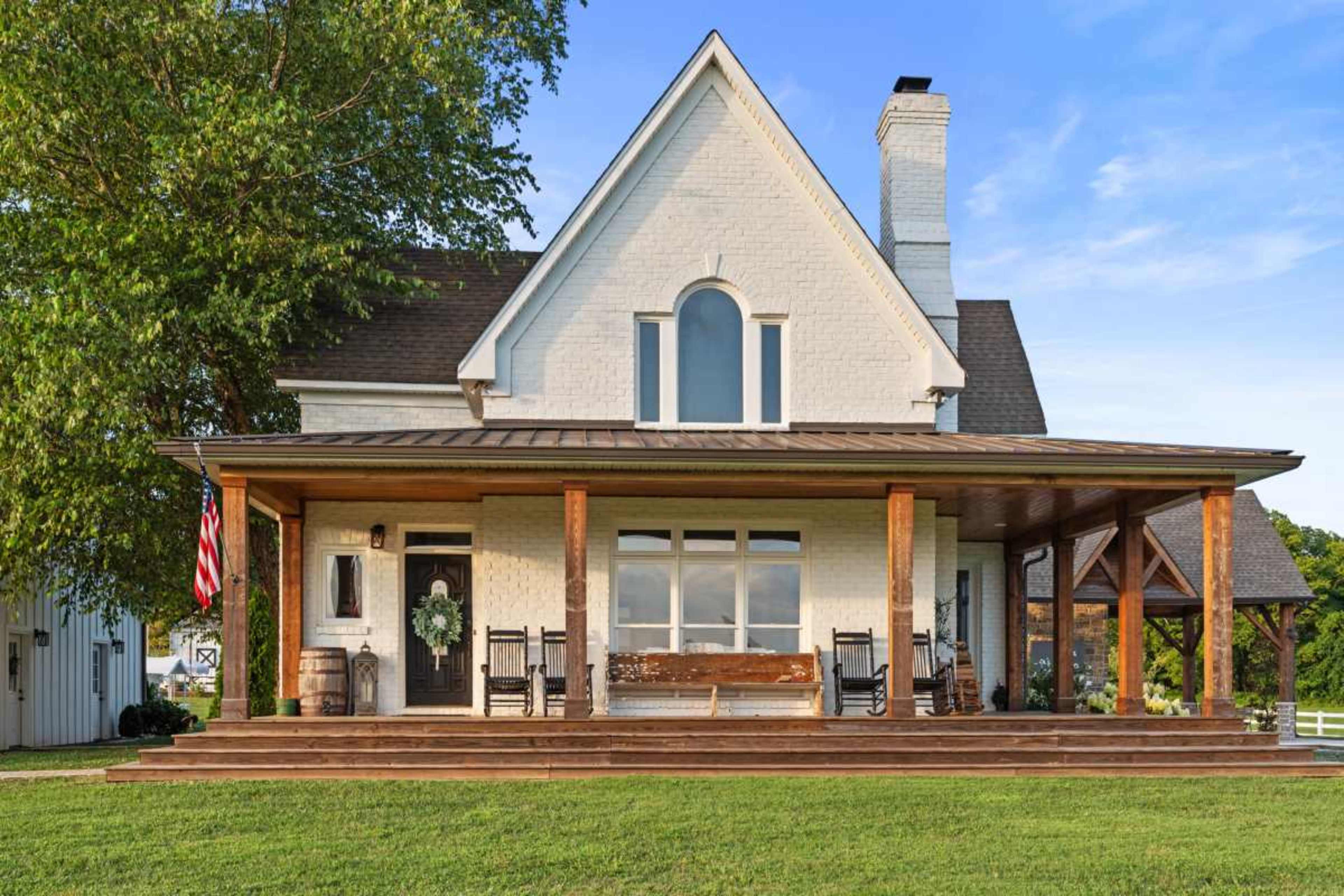 The image shows a white brick house with a large front porch, featuring wooden beams and rocking chairs, set in a green landscape under a clear blue sky.