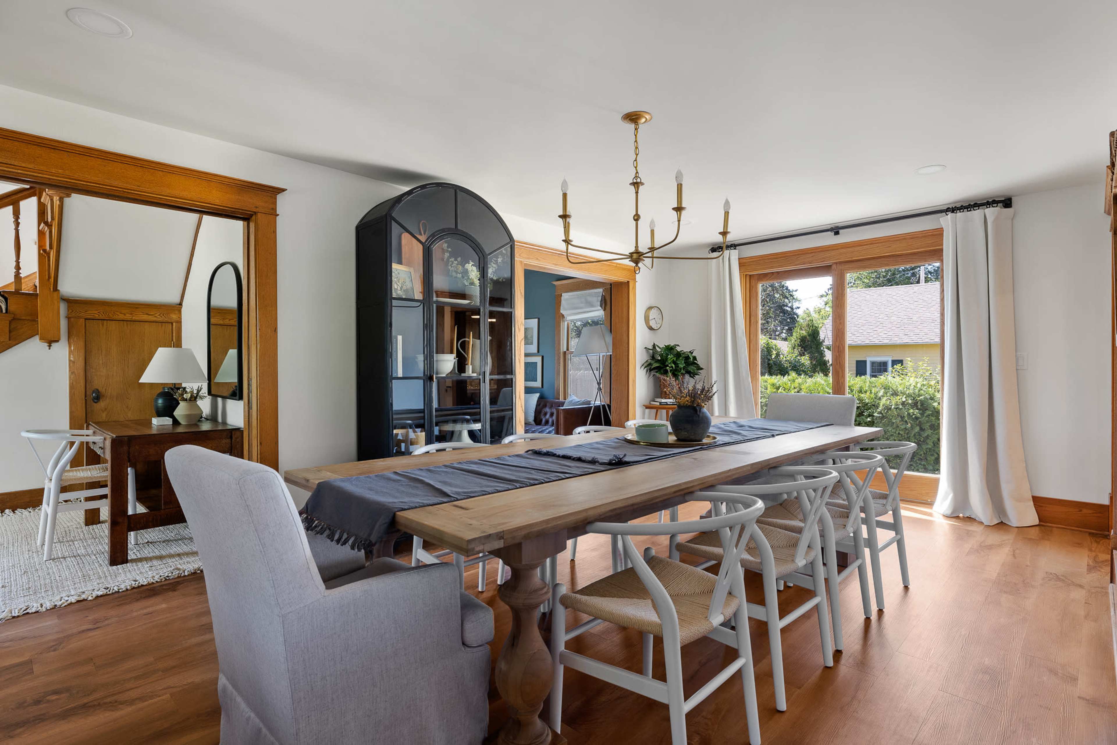 The dining room features a long wooden table surrounded by white chairs, a glass-fronted cabinet, and a chandelier, with large windows overlooking the garden.