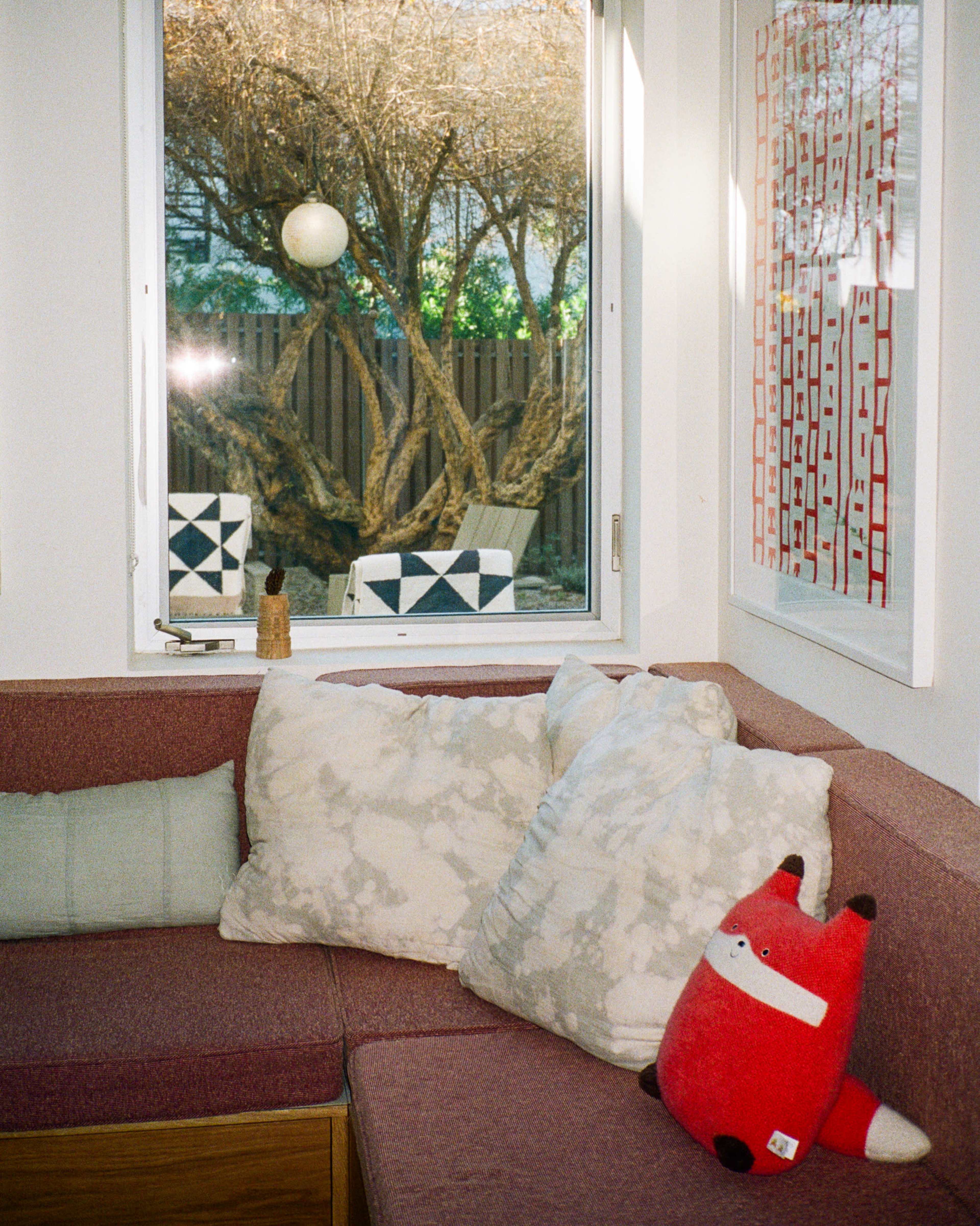 A cozy corner of a room features a sectional couch with pillows, a plush red fox toy, and a window showcasing an outdoor scene with trees and chairs.