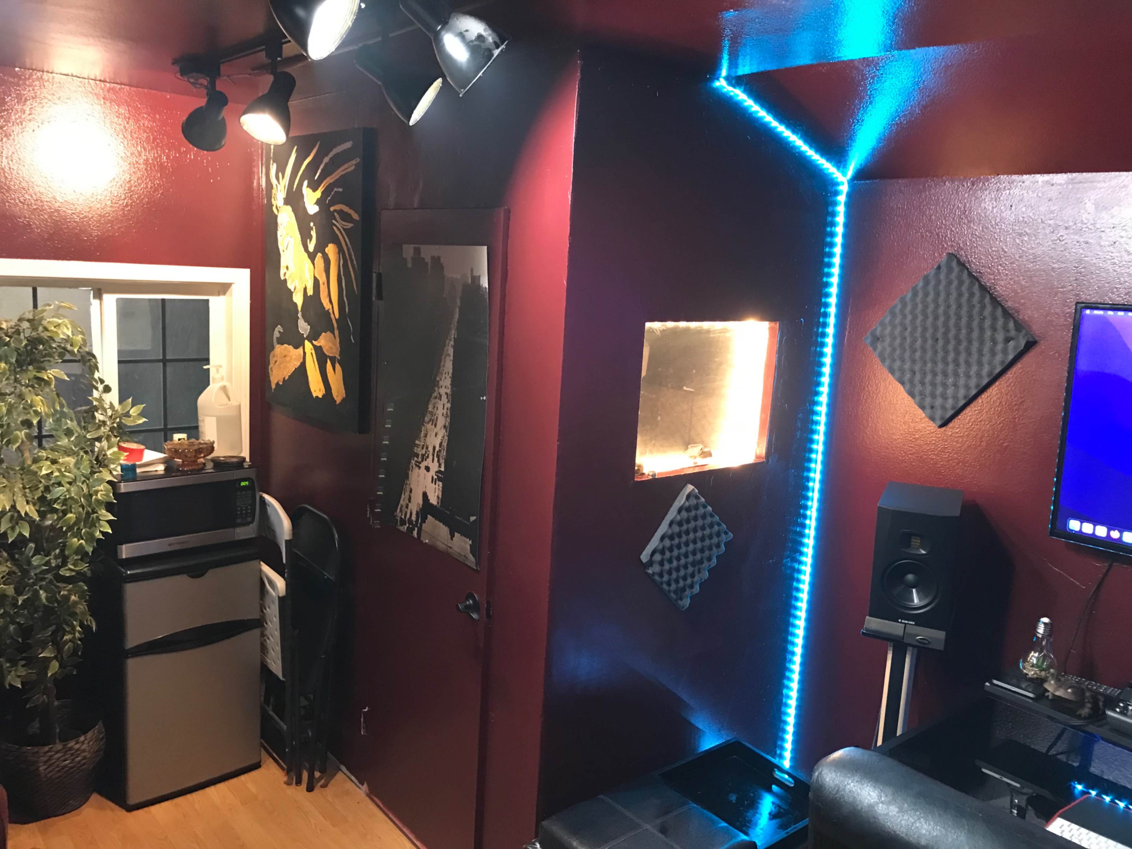 The image shows a room with maroon walls, featuring a small kitchen area with a fridge, a wall art piece, and a corner highlighted by blue LED lights.