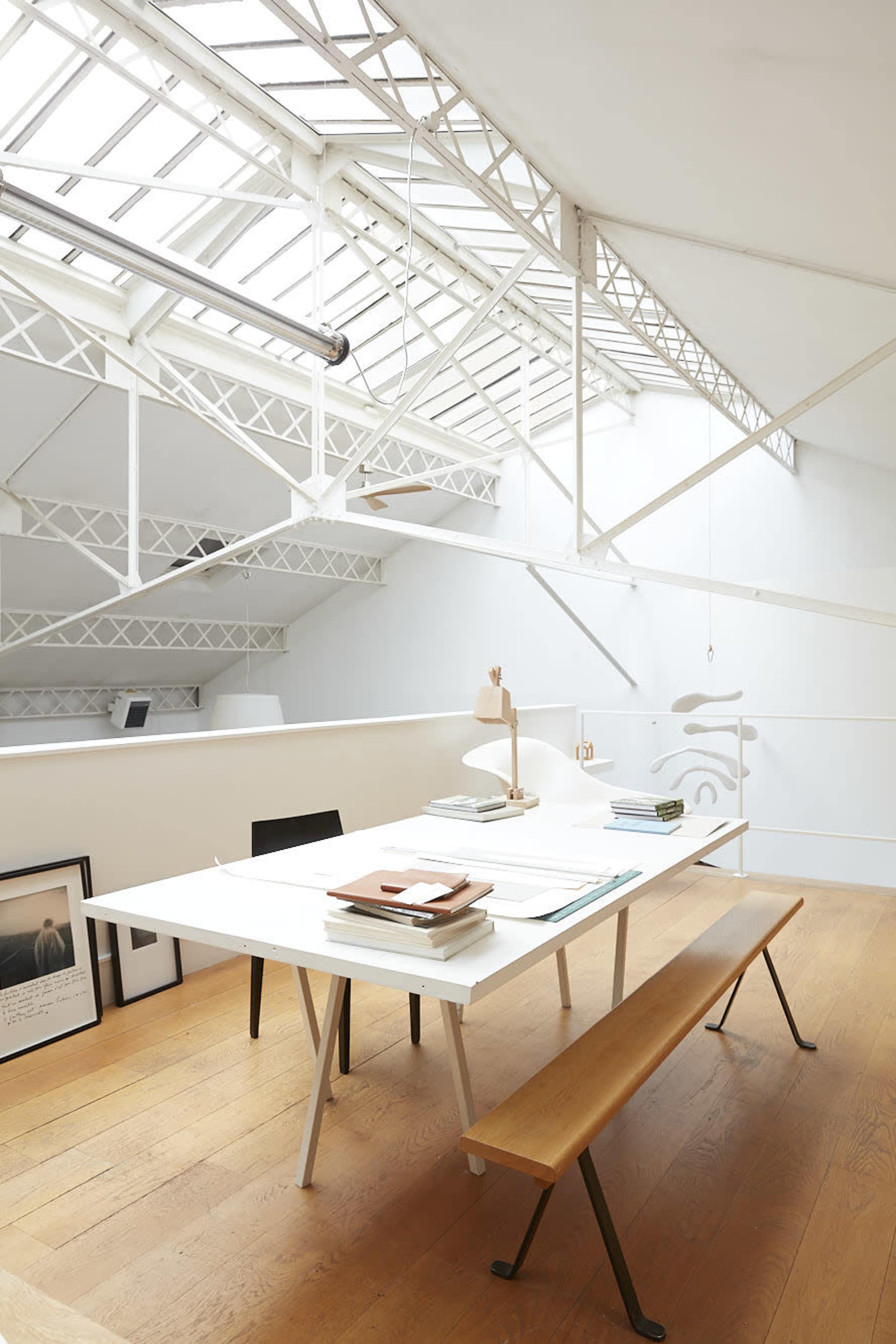 A bright, minimalist workspace features a large white table with neatly stacked materials, a lamp, and a wooden bench, all set against a backdrop of ample natural light from a skylight.