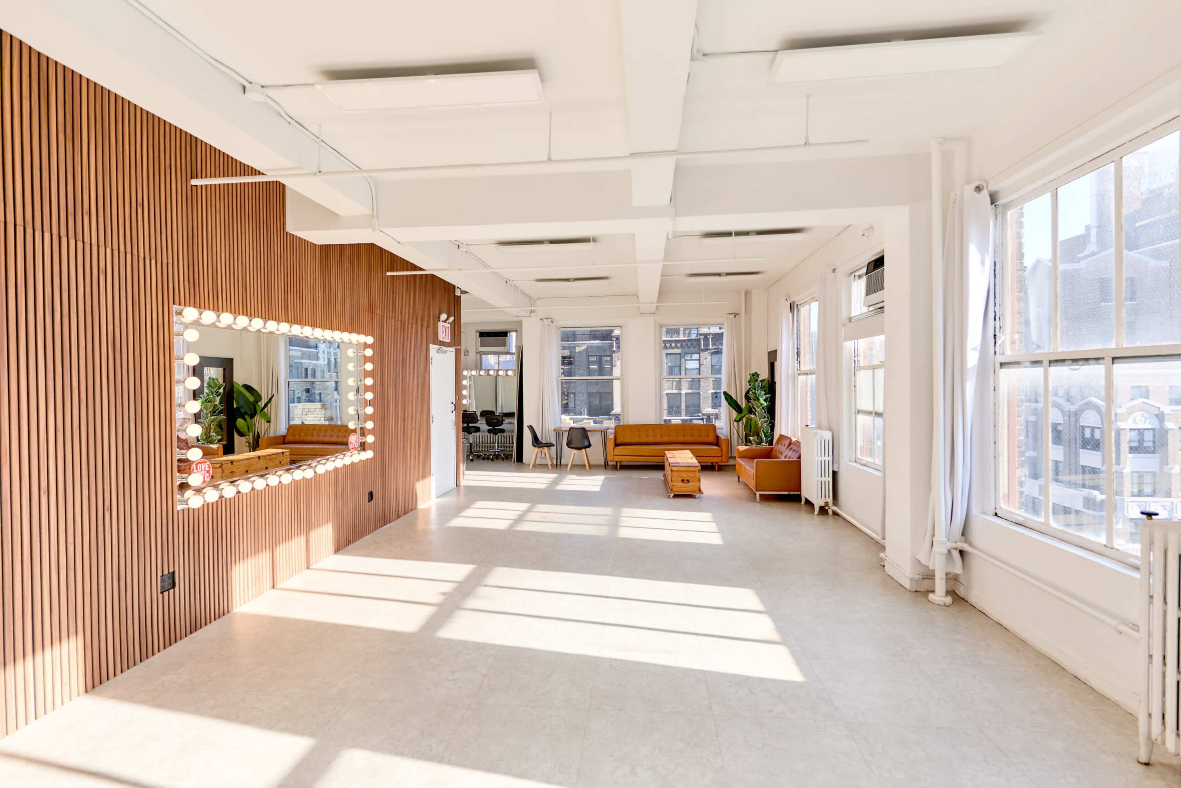 The image shows a spacious, well-lit room featuring large windows, a wooden accent wall with lighting, and a seating area with a couch.
