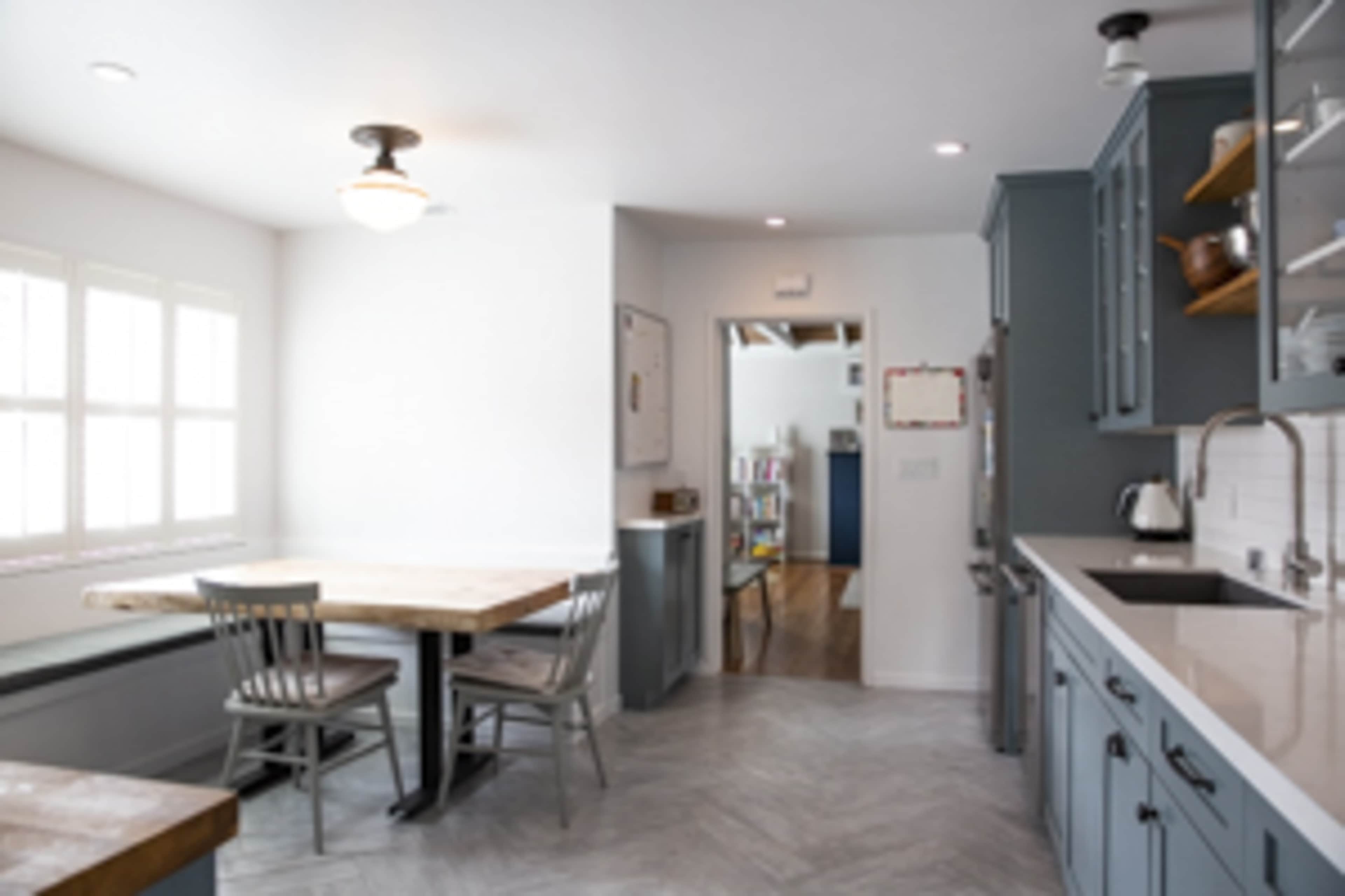 A modern kitchen features a wooden dining table with metal chairs, gray cabinets, and a doorway leading to another room.