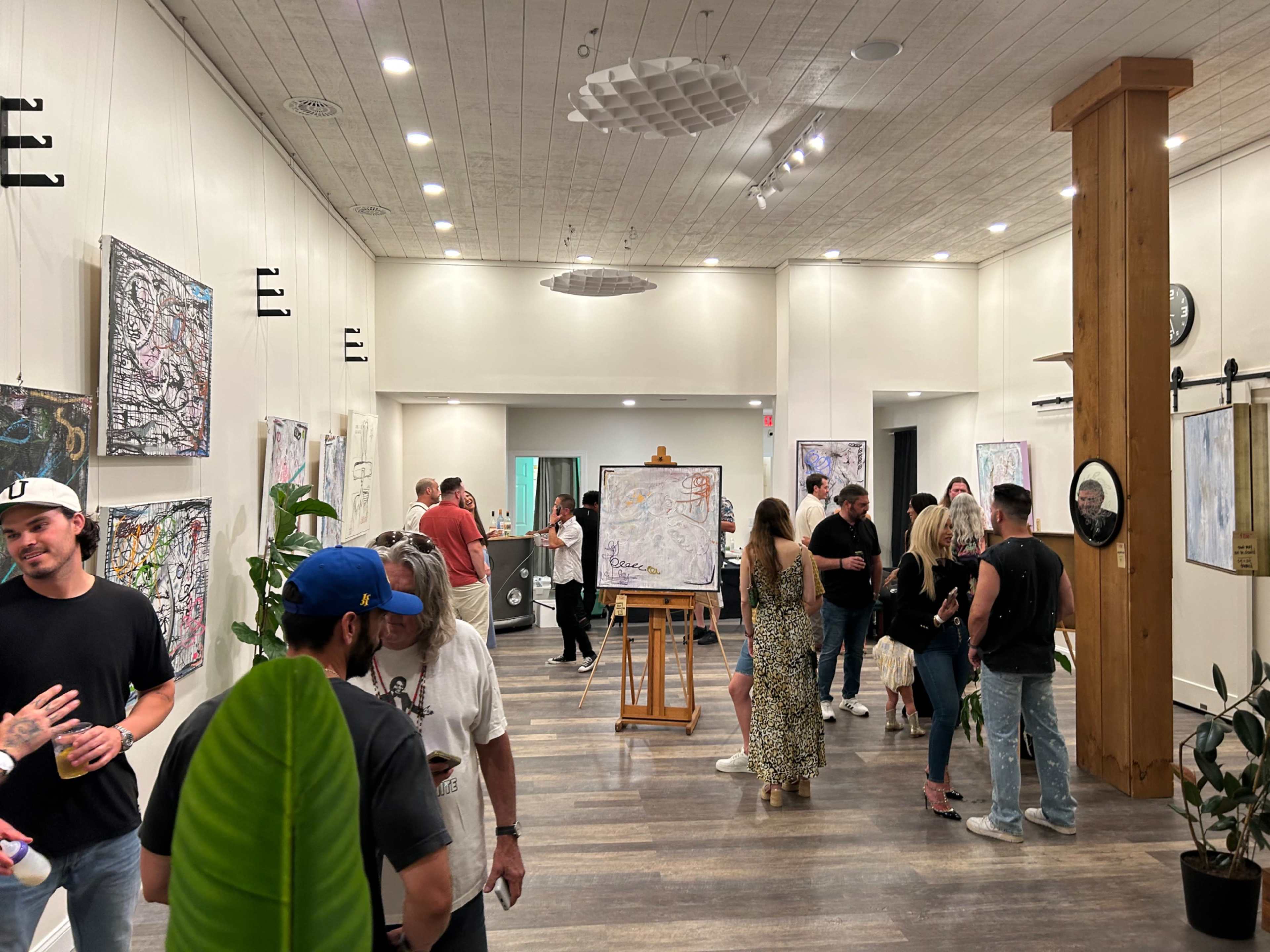 A crowded gallery features people admiring artwork displayed on white walls, with an easel holding a painting in the center.