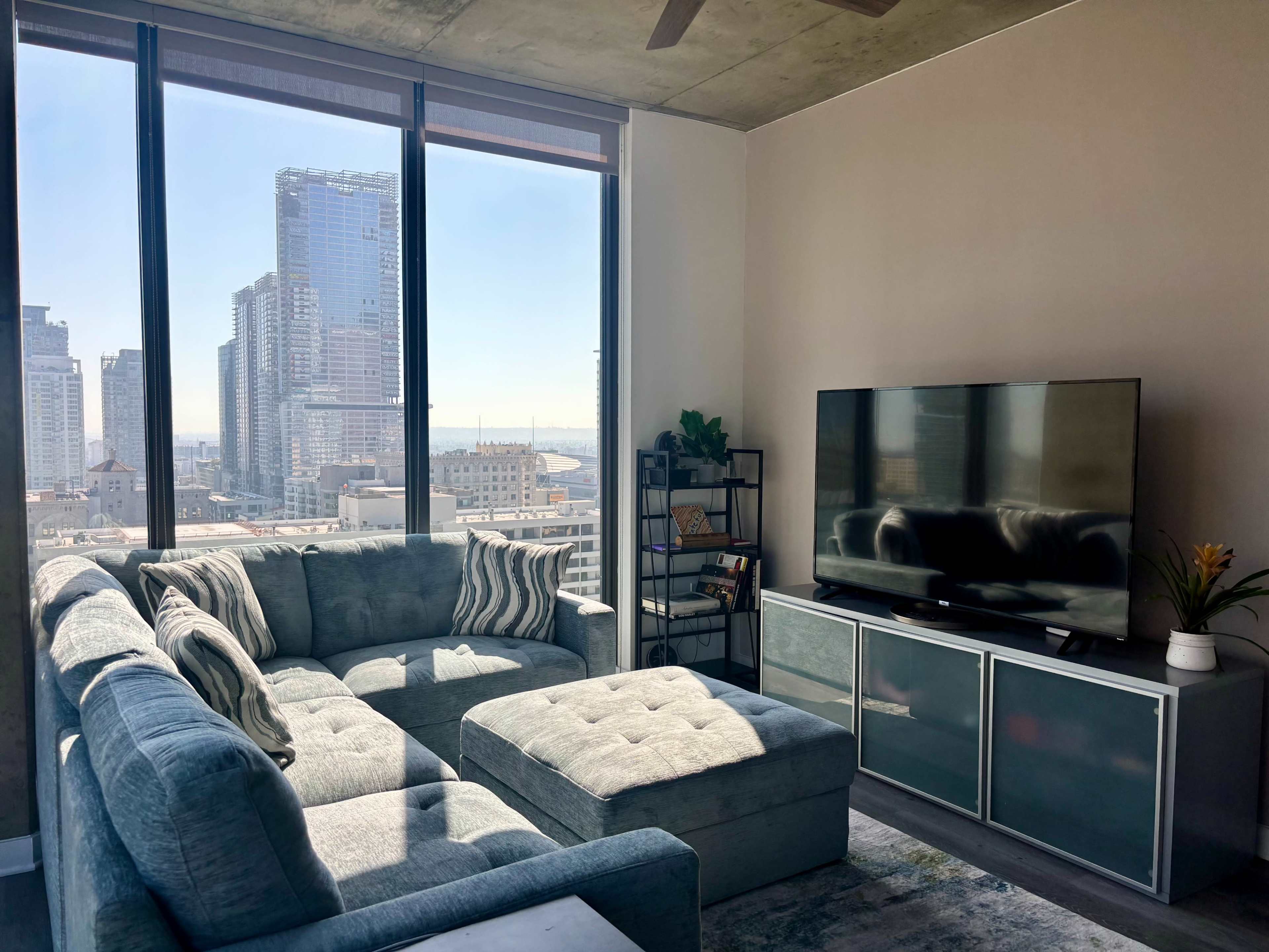 A modern living room features a large sectional couch, a television, and large windows showcasing a city skyline.