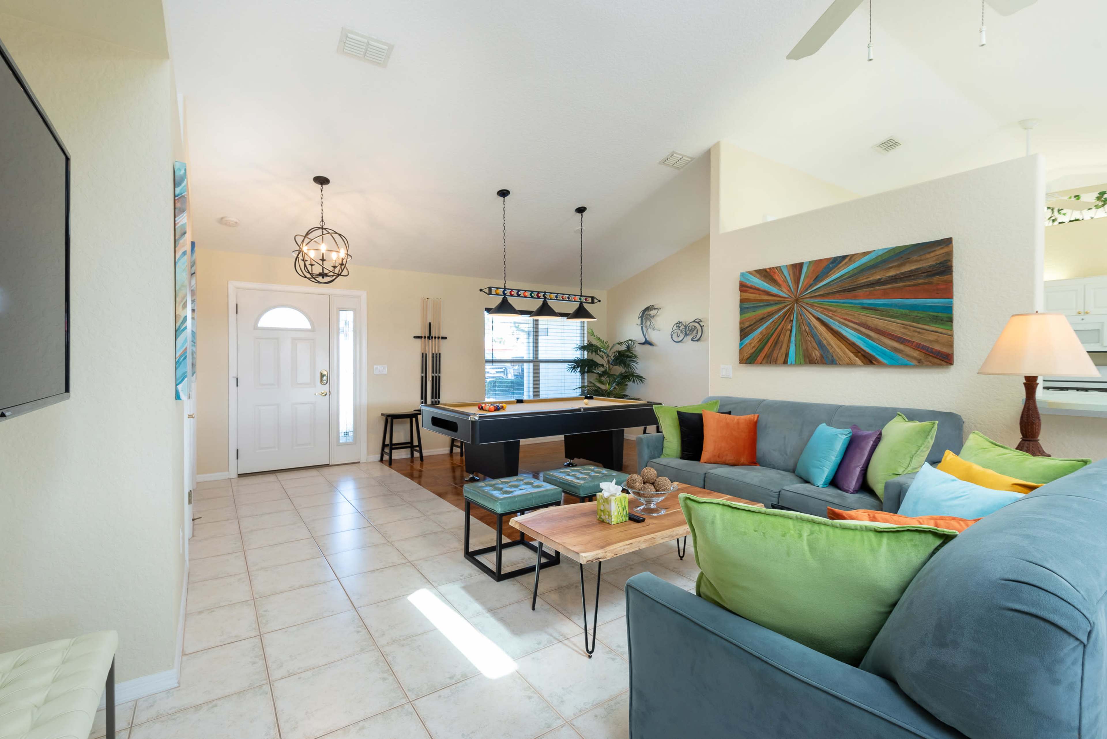 A living room with a pool table, a sectional sofa adorned with colorful cushions, and a dining area visible in the background.