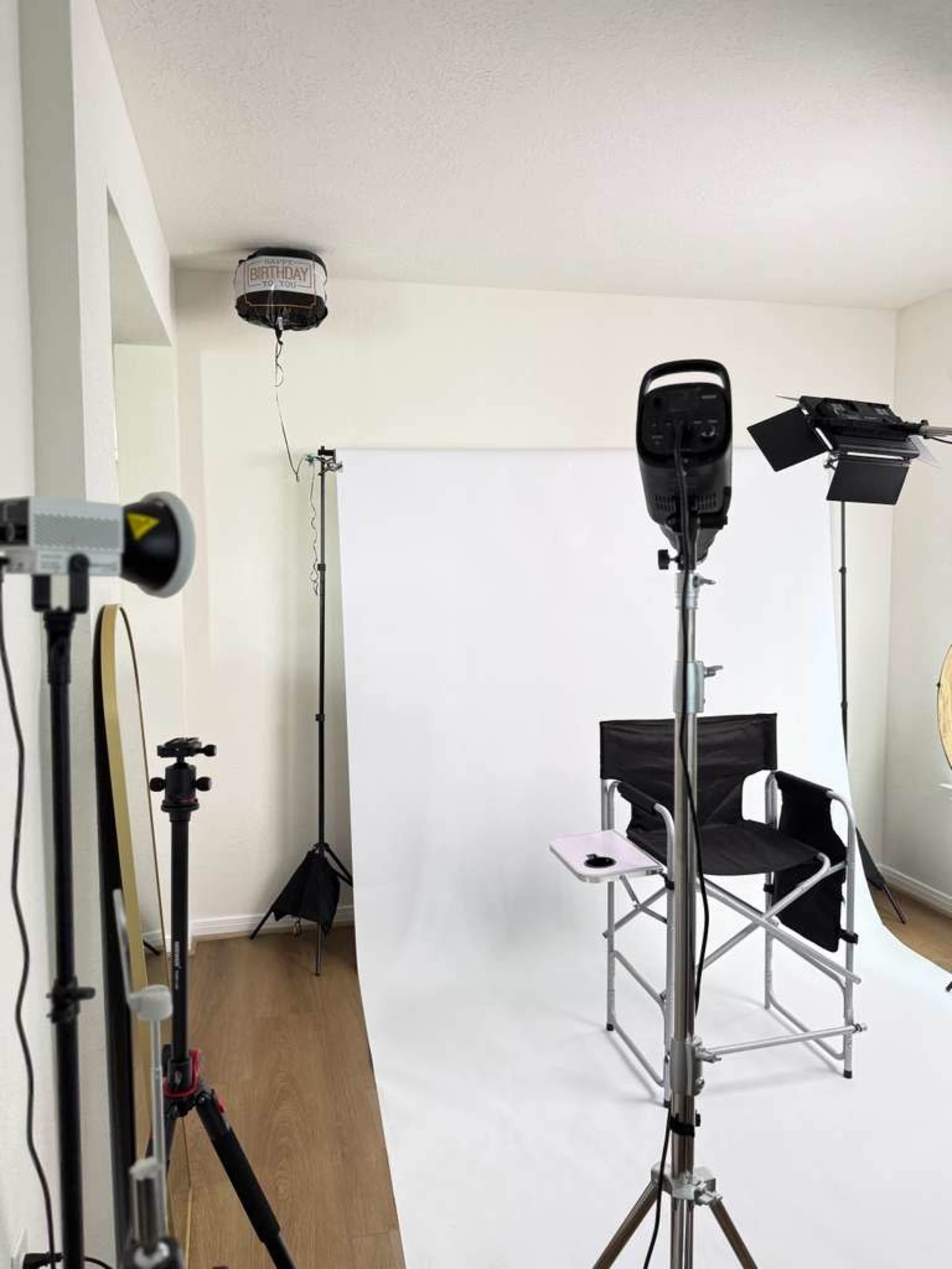 The image shows a photo studio setup with a white backdrop, a director's chair, lighting equipment, and various stands.