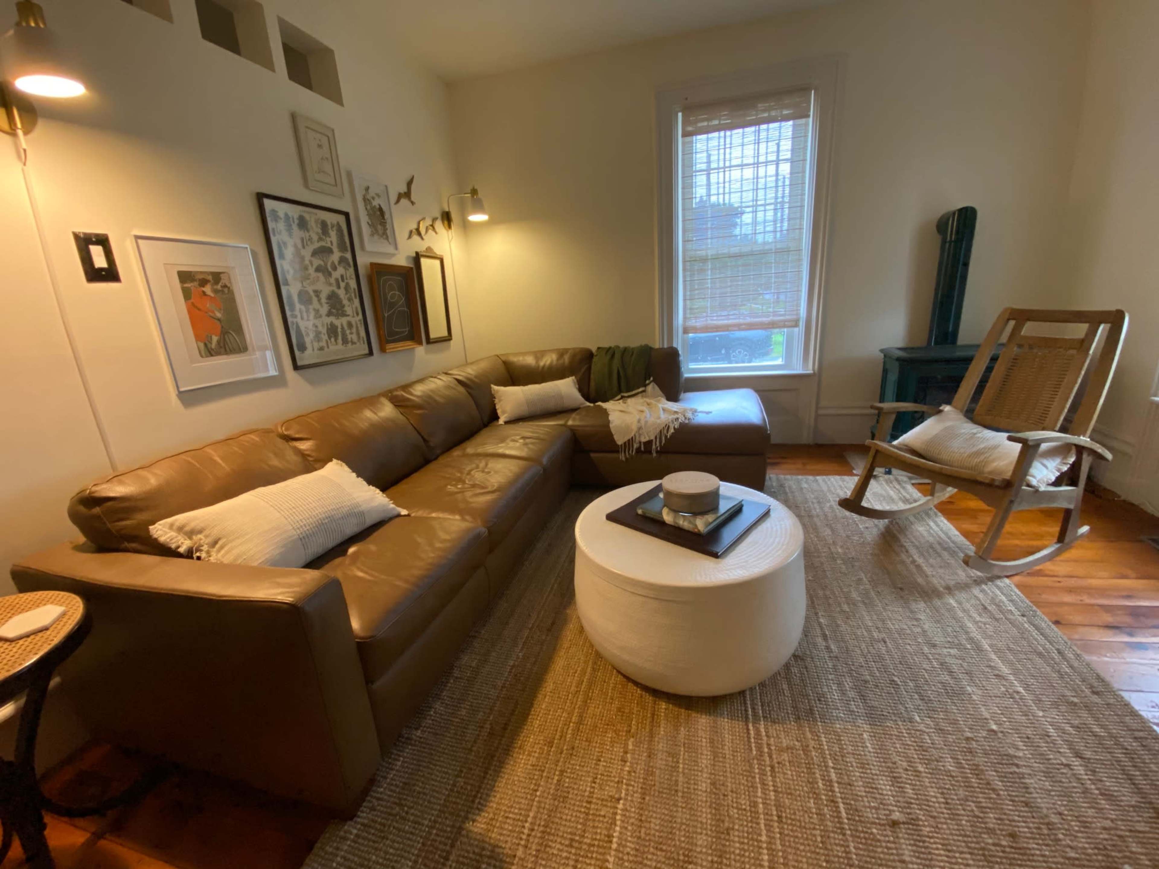 A living room with a brown sectional sofa, a round white coffee table, and a rocking chair, all arranged on a woven rug.
