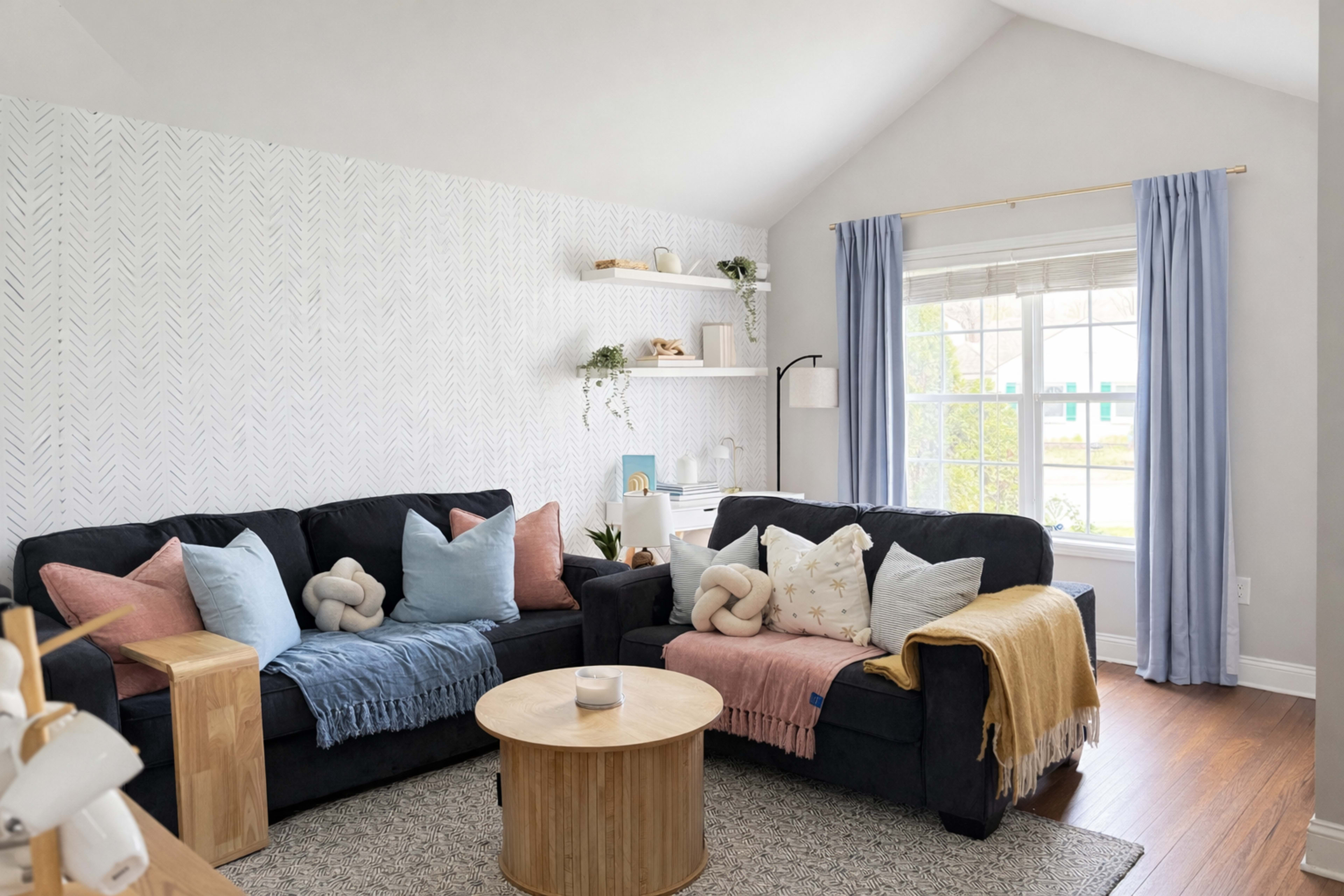 The image shows a cozy living room with two dark sofas adorned with colorful throw pillows and blankets, a round wooden coffee table in the center, and a bright window with blue curtains.
