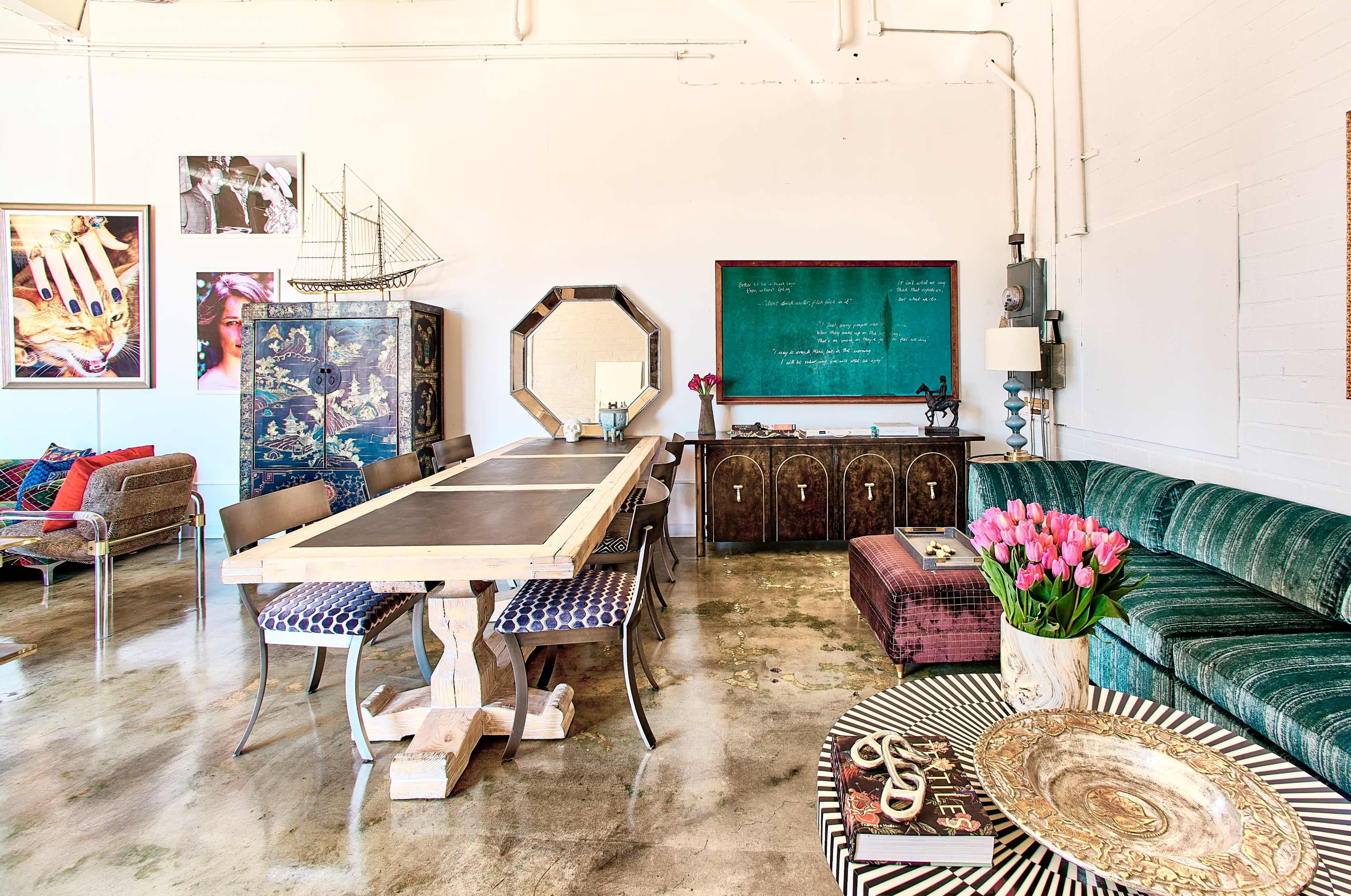The image shows a spacious interior with a large wooden dining table, several seating areas, and a chalkboard against a white wall.