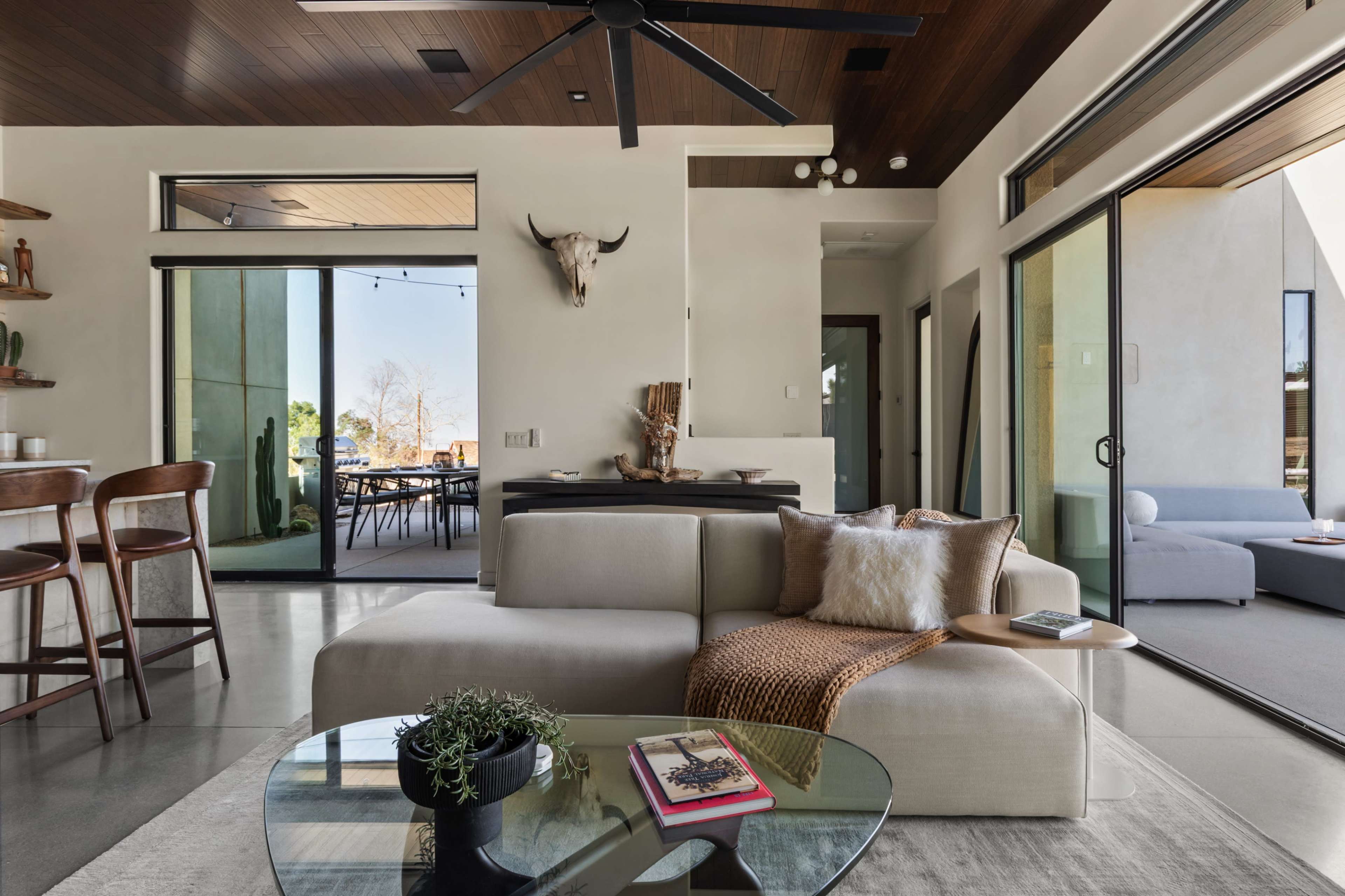 The image shows a modern living room with a beige sofa, a glass coffee table, and large windows that open to an outdoor dining area.