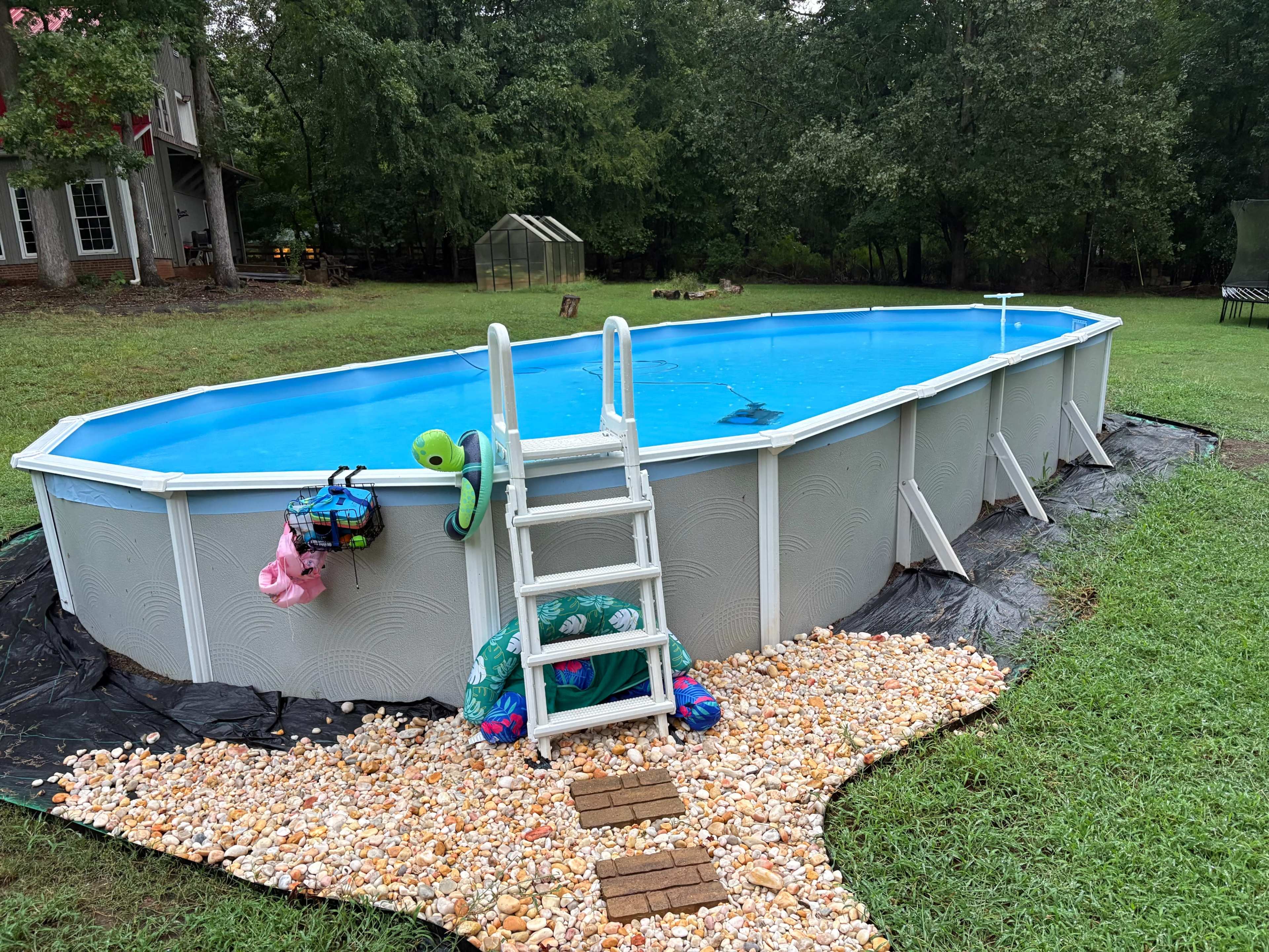An above-ground swimming pool is situated on a grassy area, surrounded by a decorative pebble border and near a green structure in the background.