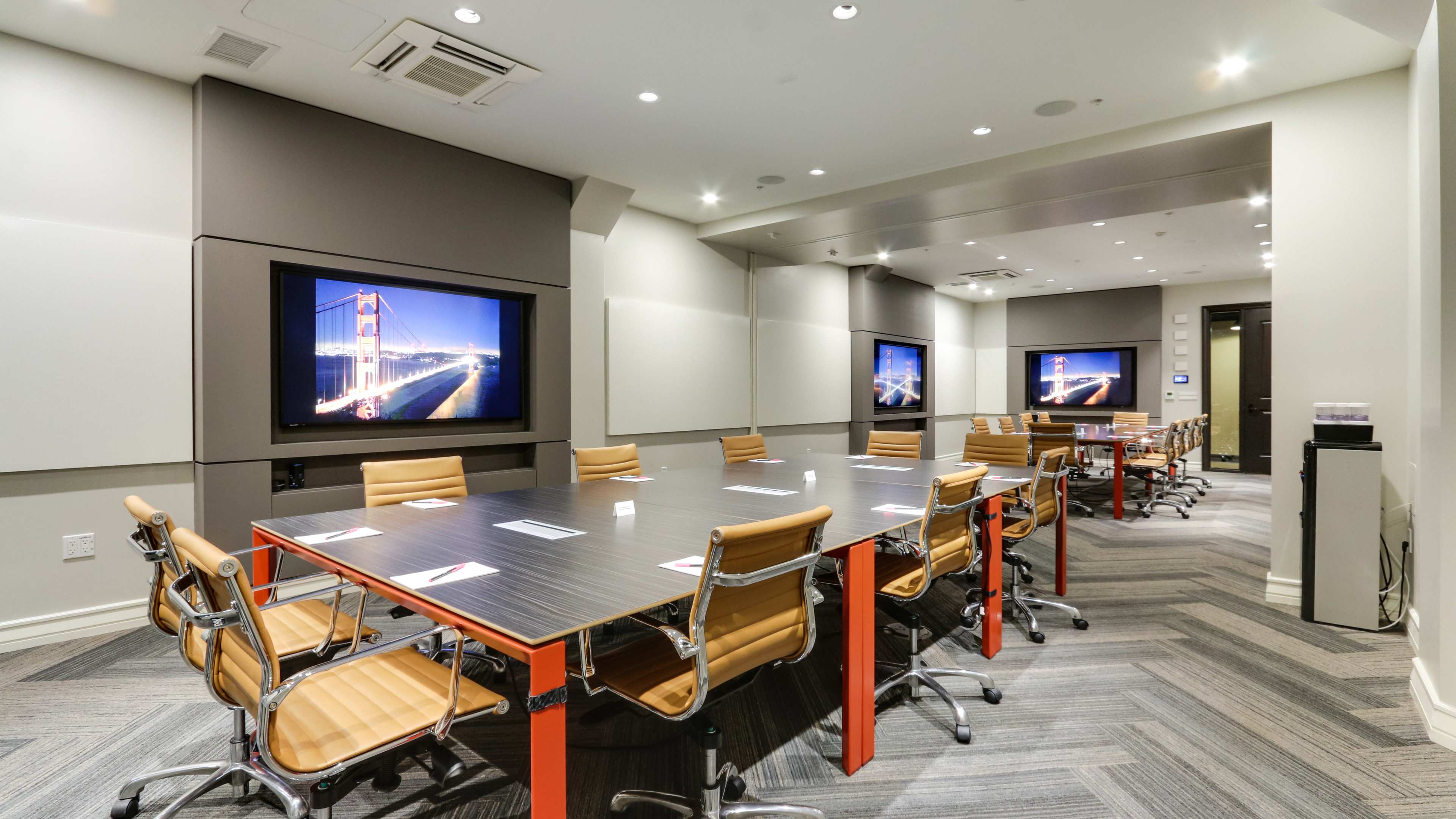 The image shows a modern conference room equipped with a large table, ergonomic chairs, and two wall-mounted screens displaying an image of the Golden Gate Bridge.