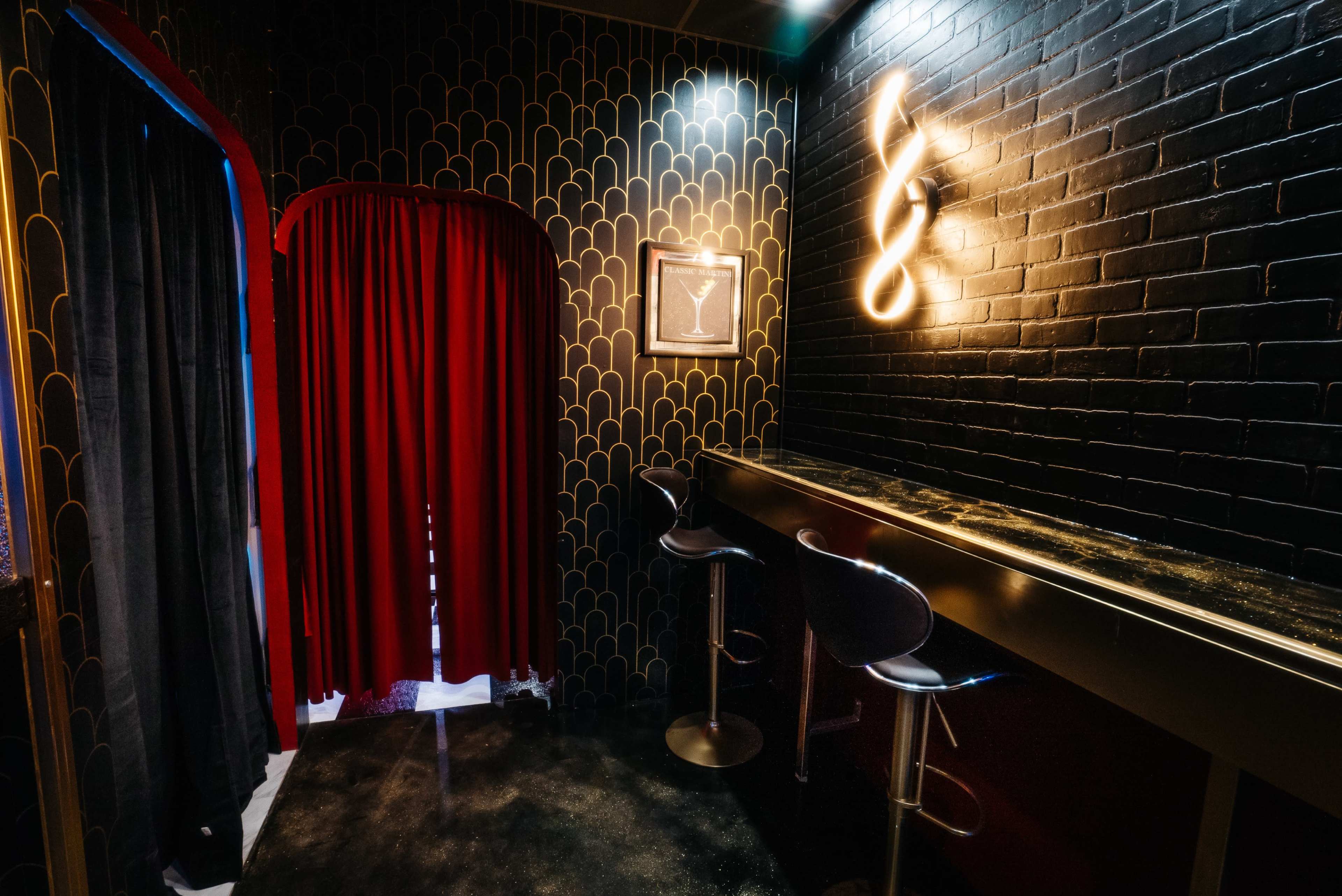 A bar area features a dark brick wall, a glowing light fixture, a martini-themed framed artwork, and a red curtain leading to another space.