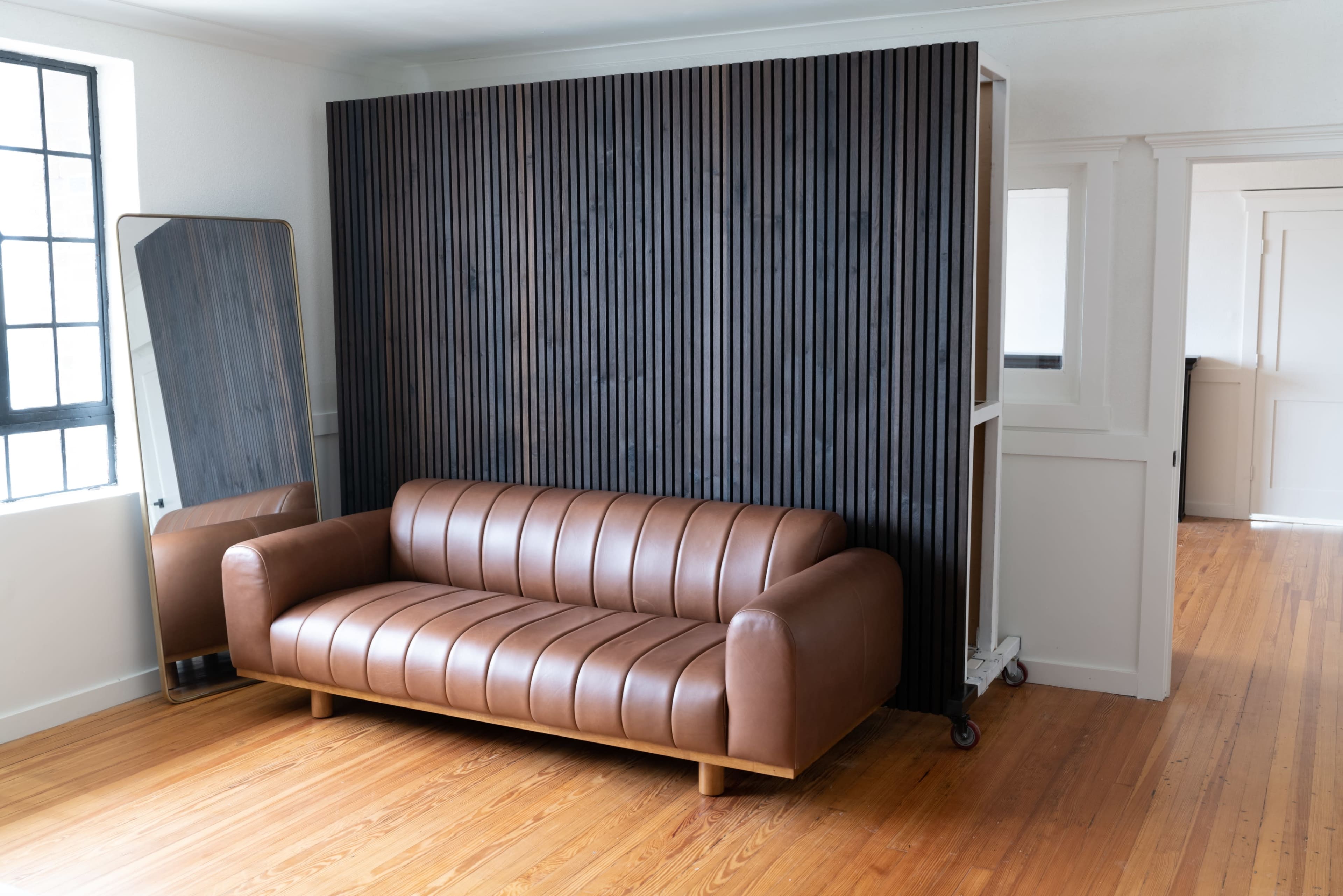 A brown leather sofa is positioned against a vertical slatted wood wall in a minimalist room with hardwood flooring.