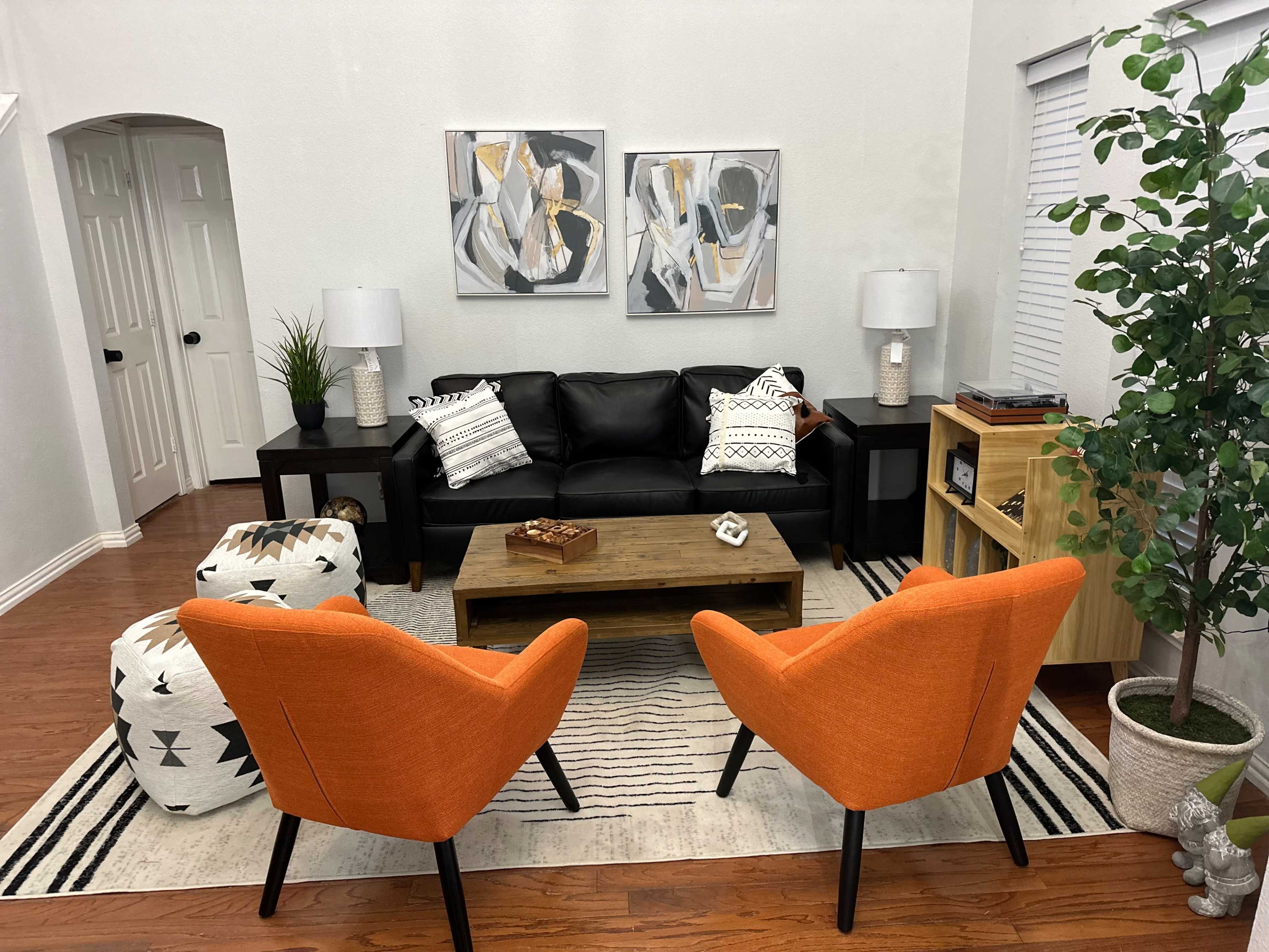 A modern living room features a black leather sofa, two orange chairs, a wooden coffee table, and abstract artwork on the walls.
