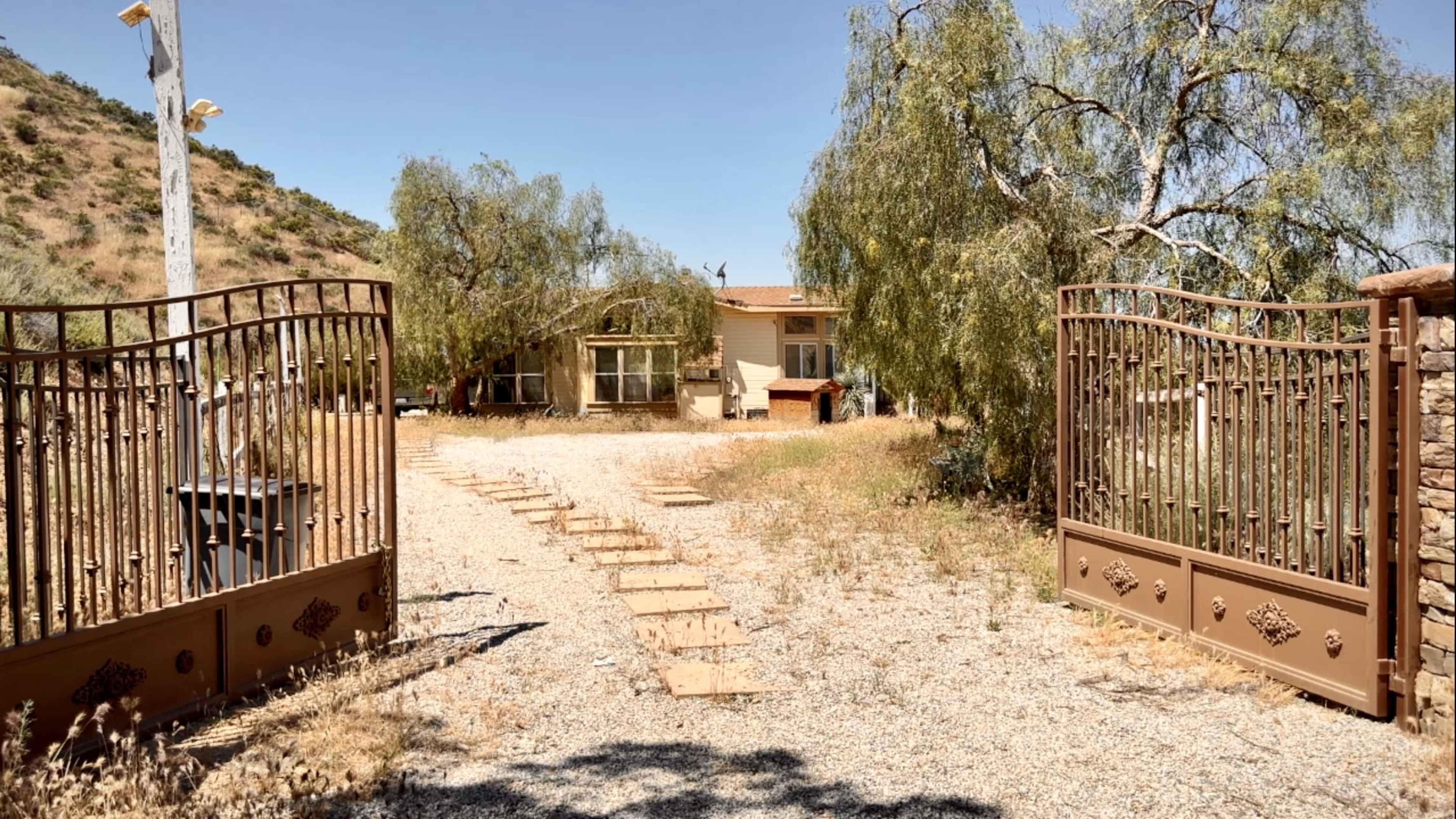 A gated entrance leads to a house surrounded by trees and gravel on a sunny day.