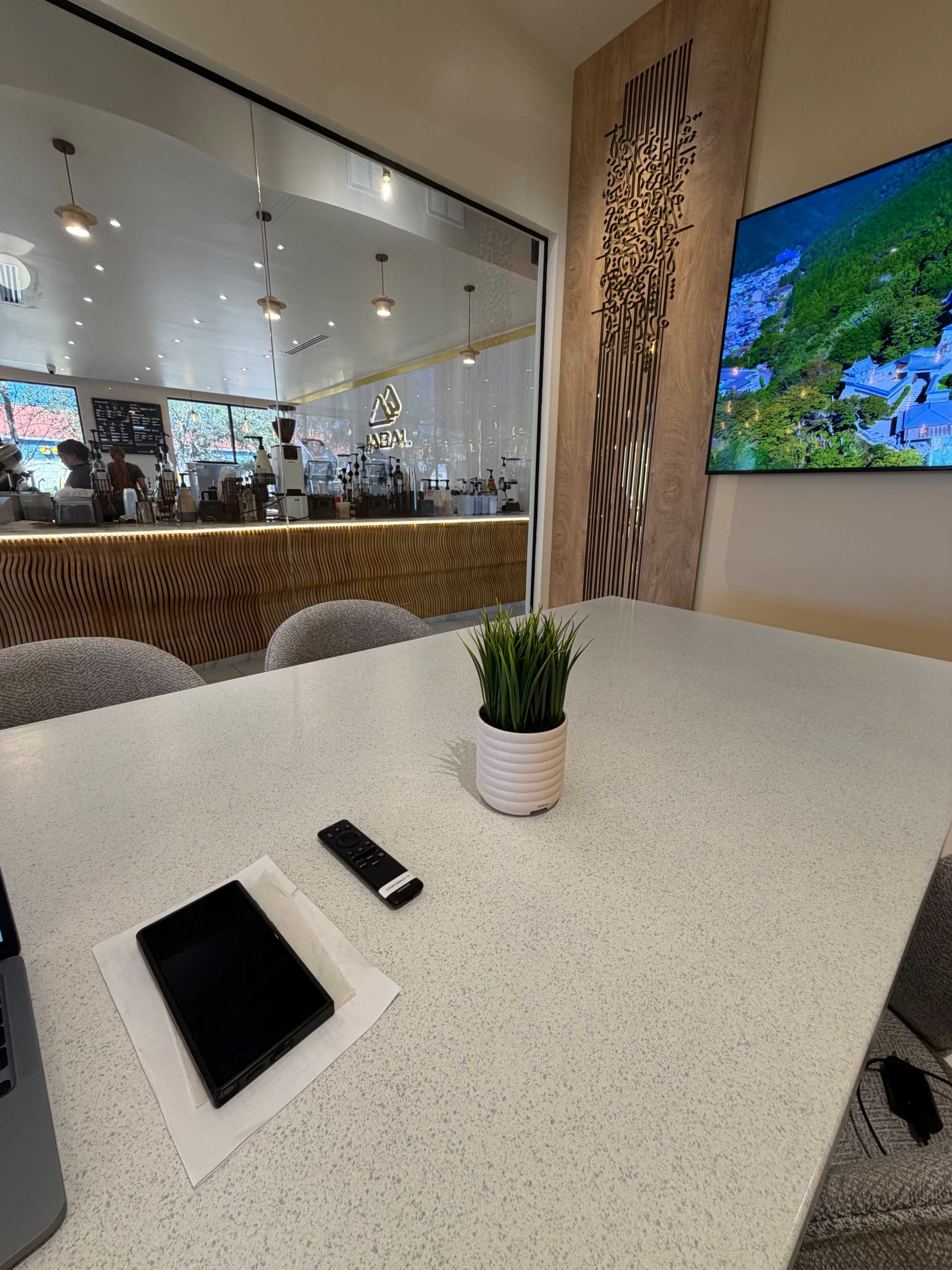 The image shows a table with a small plant, a remote control, and a tablet, while reflecting a cafe setting visible through glass walls.