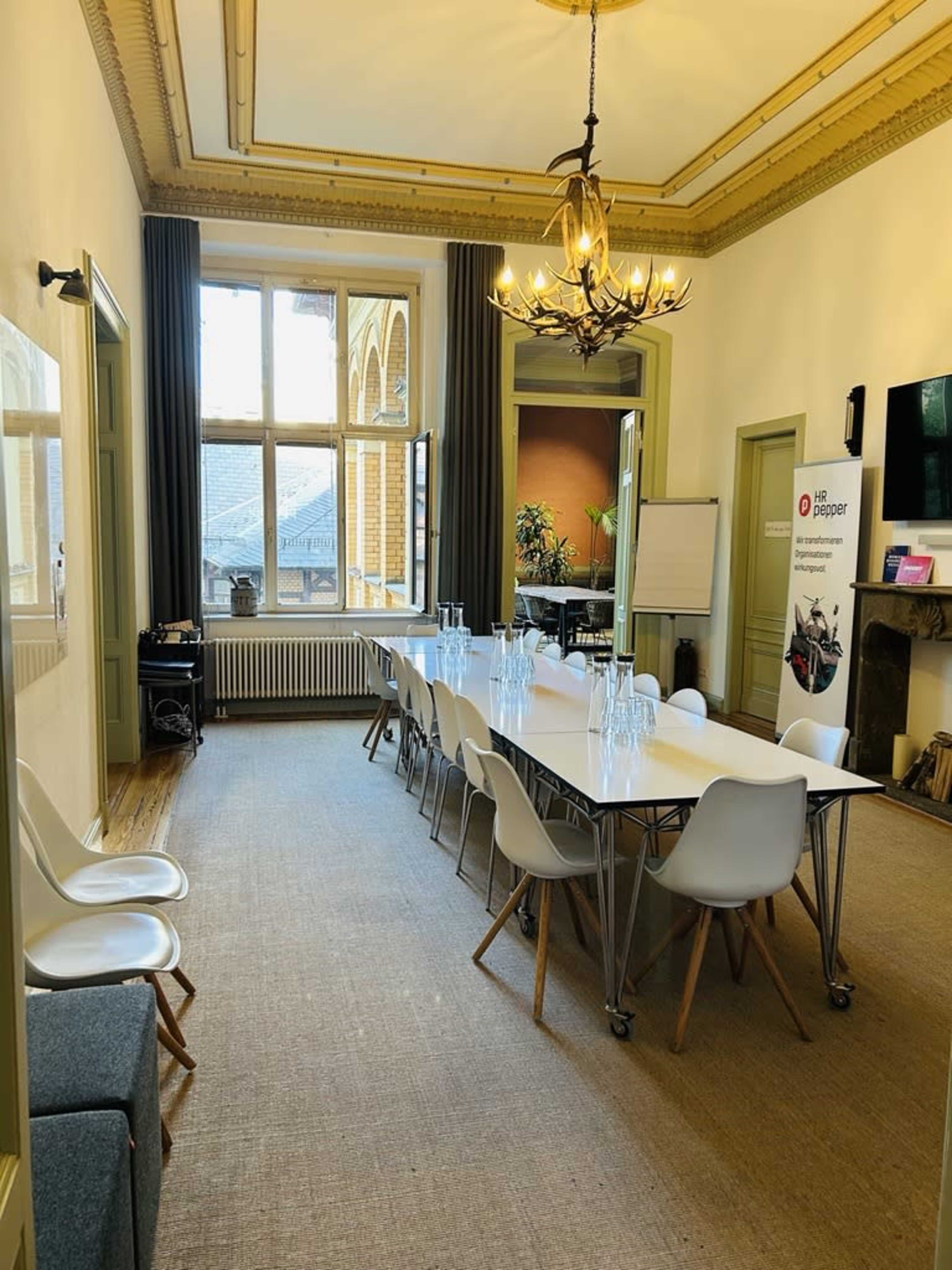 The image shows a spacious meeting room with a long table set for a presentation, featuring modern chairs, large windows, and decorative light fixtures.