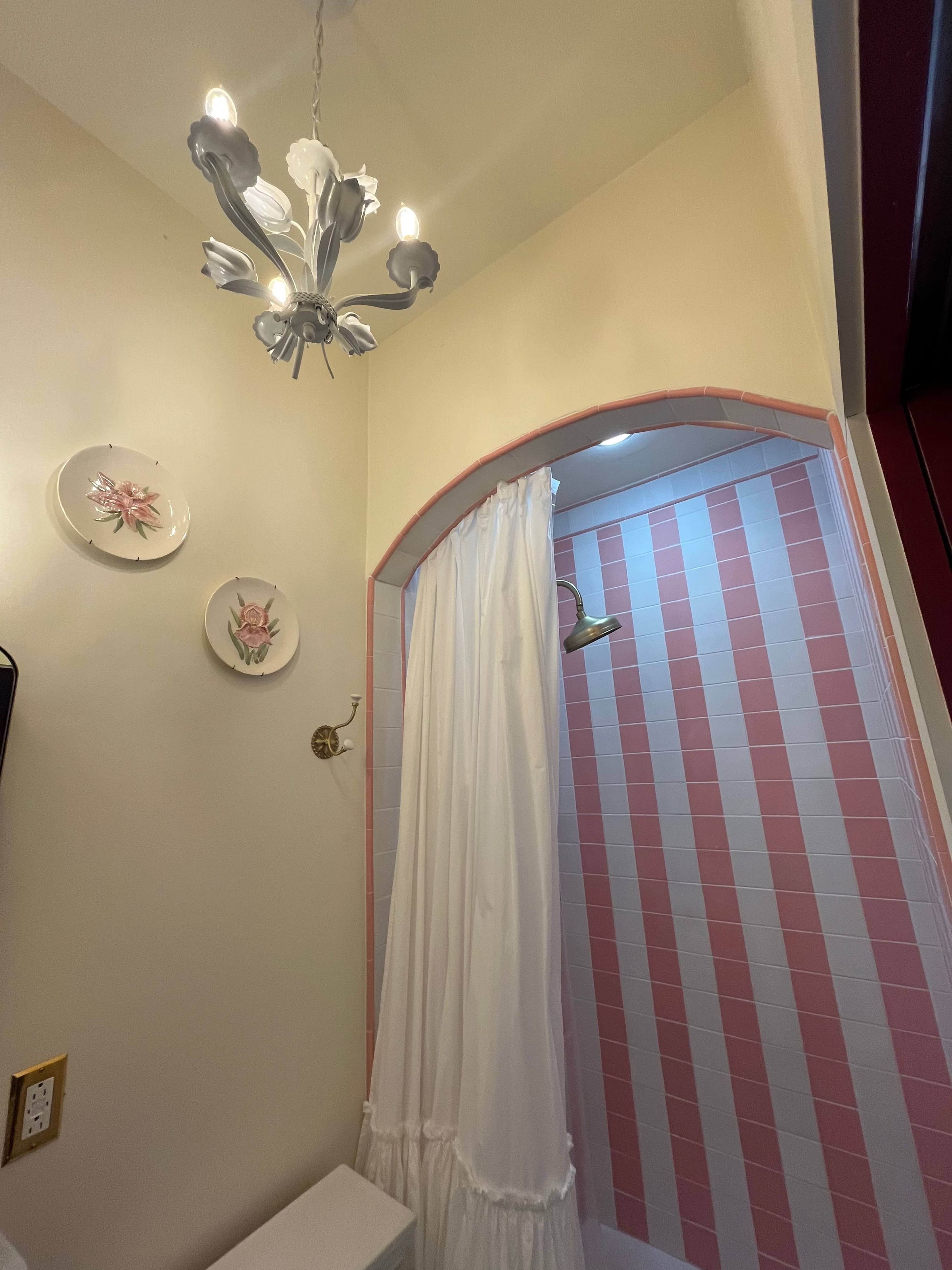 The image shows a small bathroom shower area with a pink and white tiled wall, a white curtain, and a chandelier above.