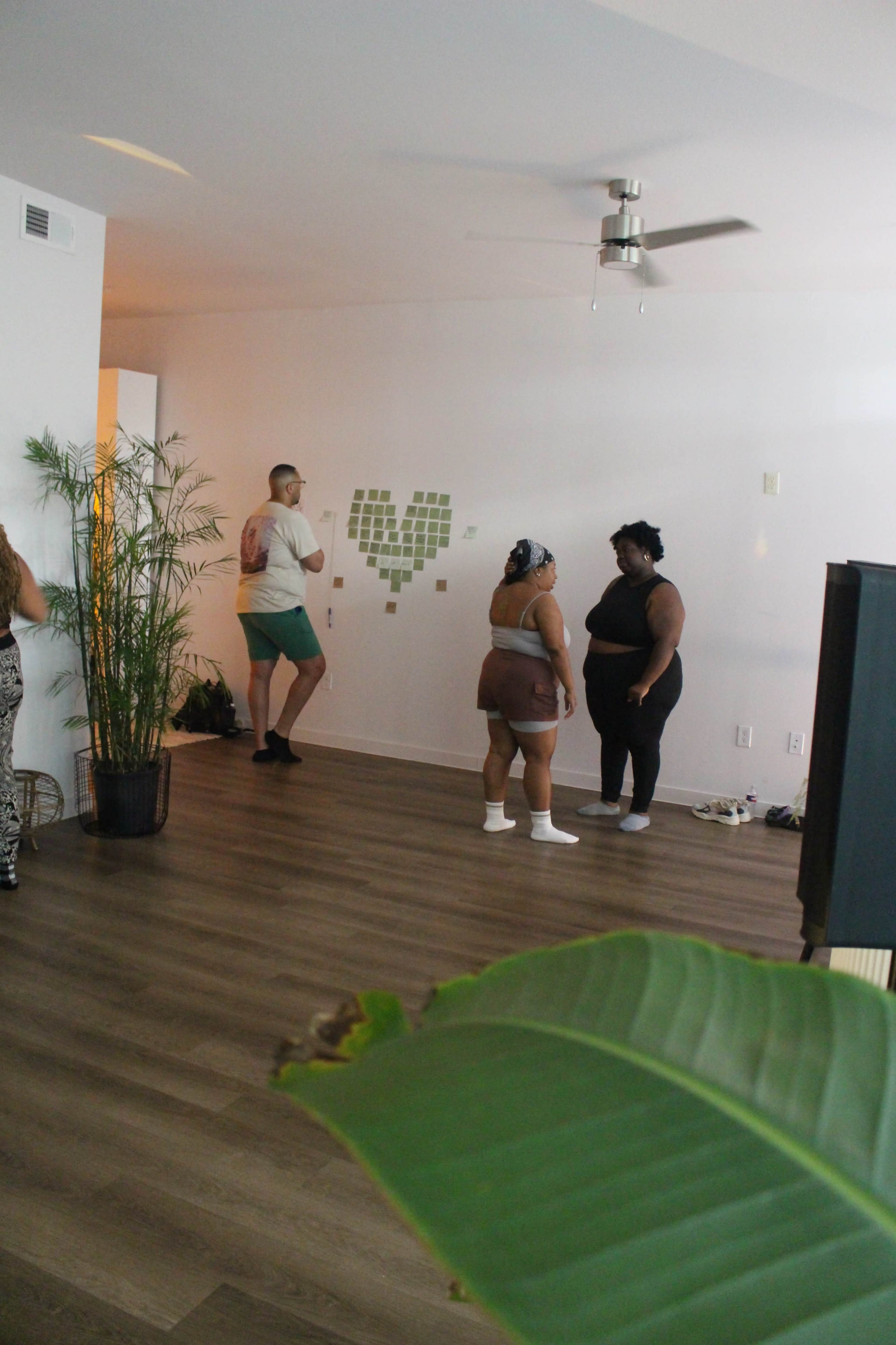 Three people interact in a modern, softly lit room, with a green plant in the foreground and a wall decorated with arranged green tiles.