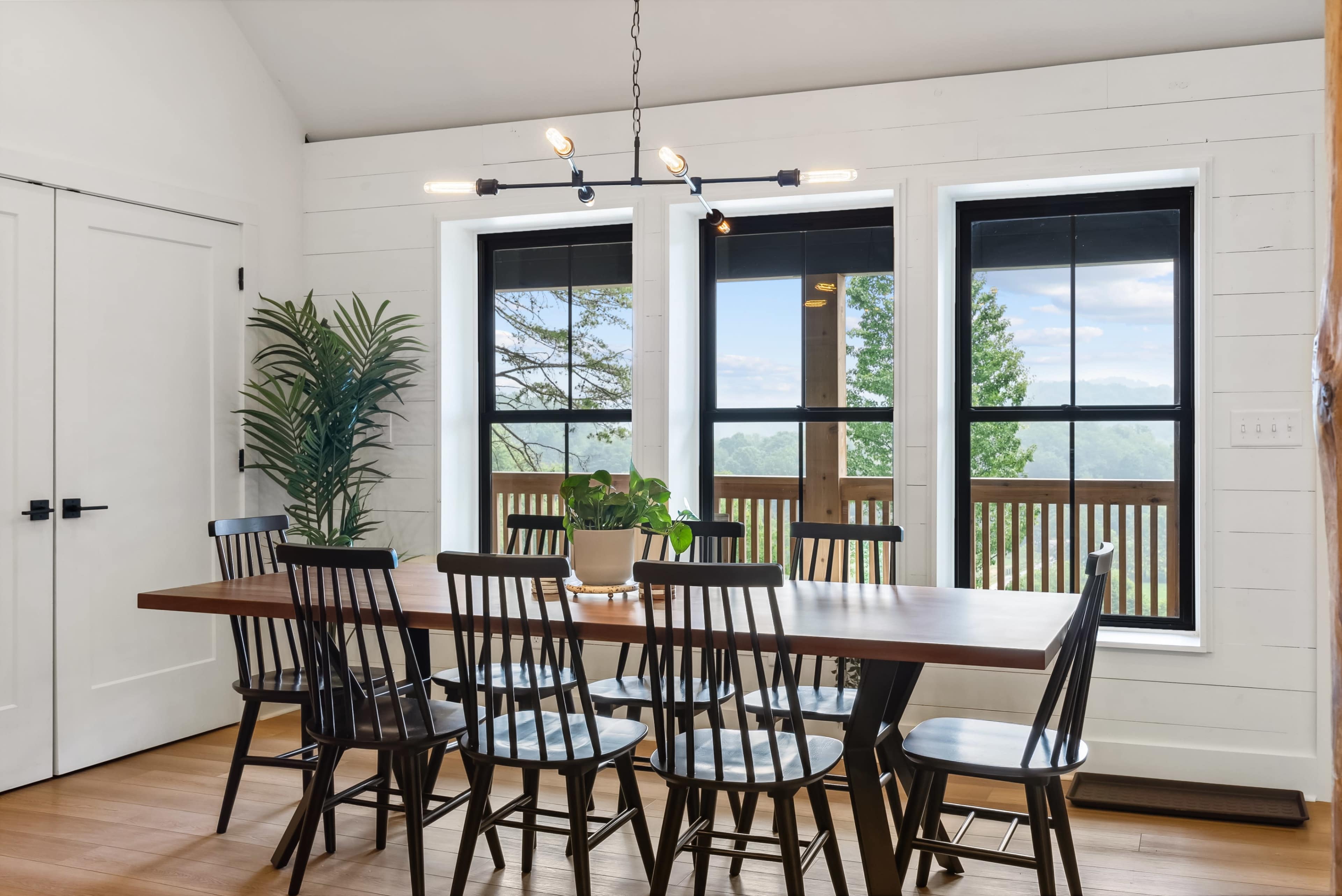 A dining room features a large wooden table surrounded by black chairs, with large windows offering a view of trees and mountains.