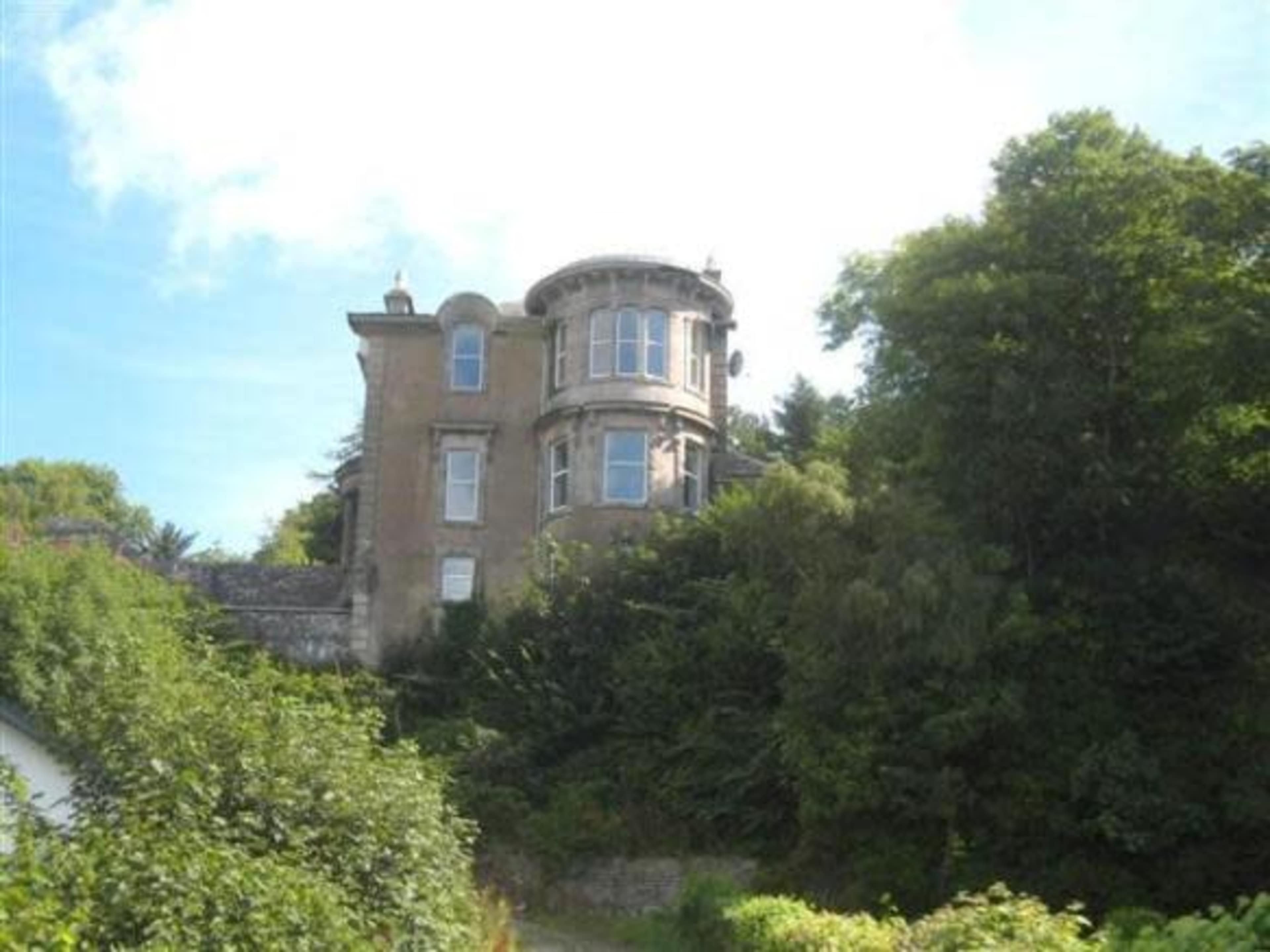 A large, historic house with a circular tower is set against a backdrop of greenery.