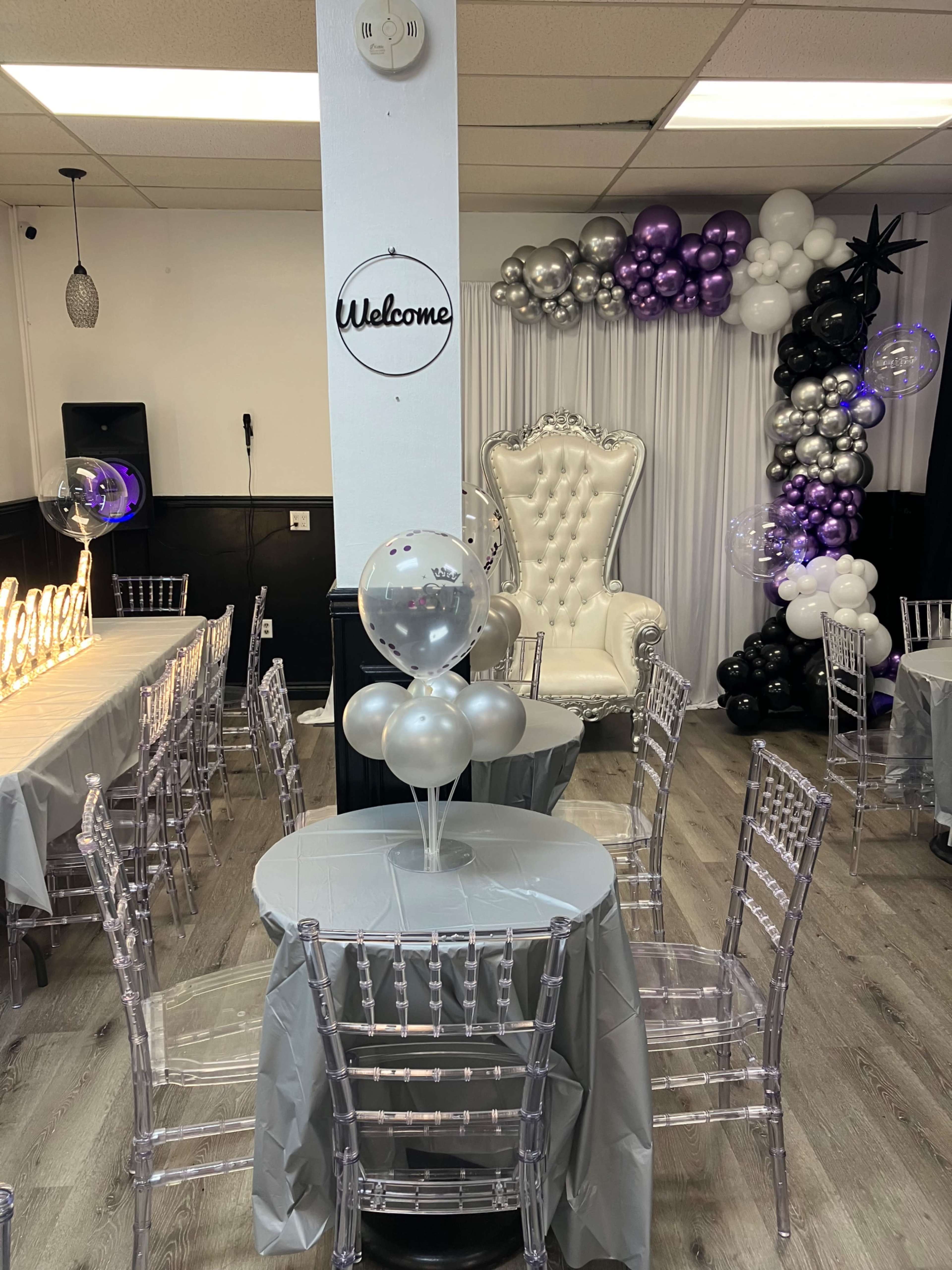 The image shows an event space decorated with silver and purple balloons, a white throne chair, and tables covered with gray tablecloths.